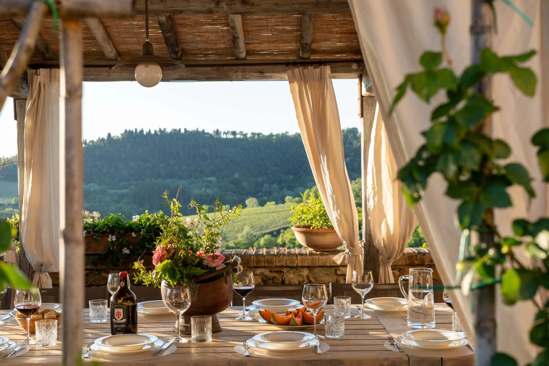 A rustic outdoor dining area with a wooden table set with white plates, glasses, and a bottle of red wine, surrounded by greenery and hills in the background.