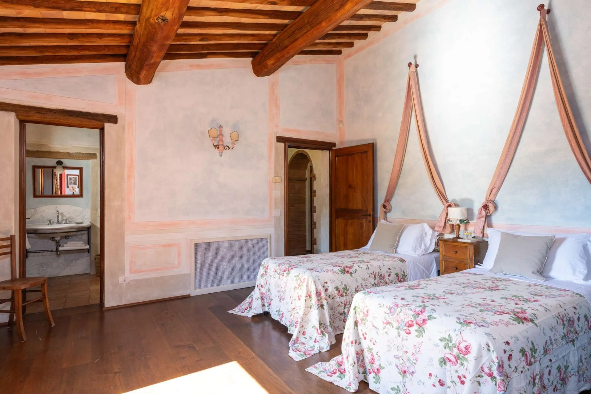 A cozy bedroom with two twin beds covered in floral bedspreads, pink canopy drapes, wooden nightstands, and a rustic ceiling with exposed beams. A small mirror hangs on the wall, and a doorway reveals a bathroom with a sink and a mirror.
