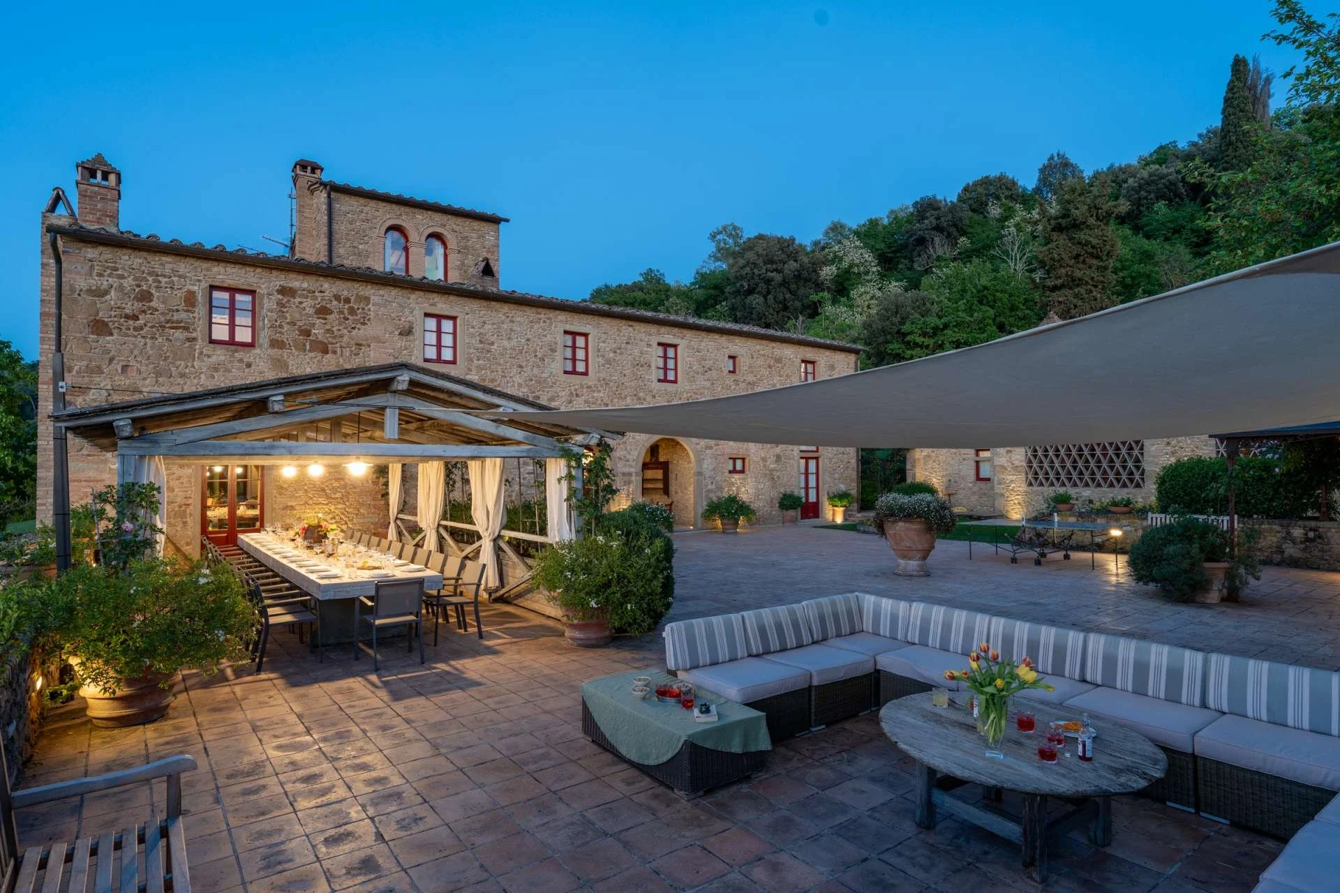 An outdoor patio area at dusk with string lights, a long dining table with chairs under a canopy, a striped outdoor sofa, and a stone house with multiple windows and a hill with trees in the background.