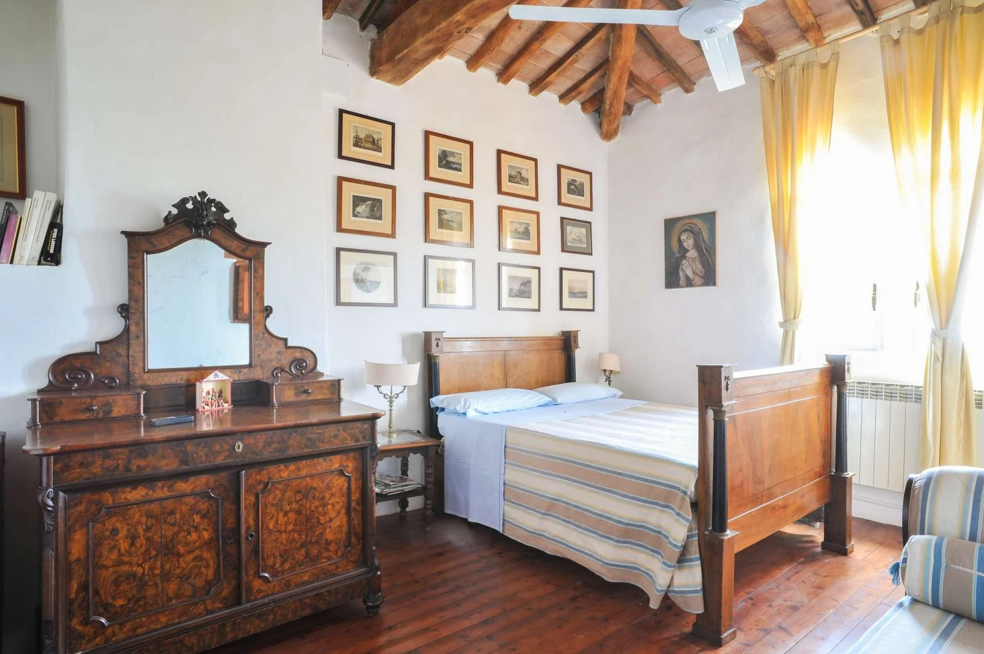 A vintage bedroom with a wooden bed, dresser, and wall art, featuring a ceiling fan, window with yellow curtains, and hardwood floors.