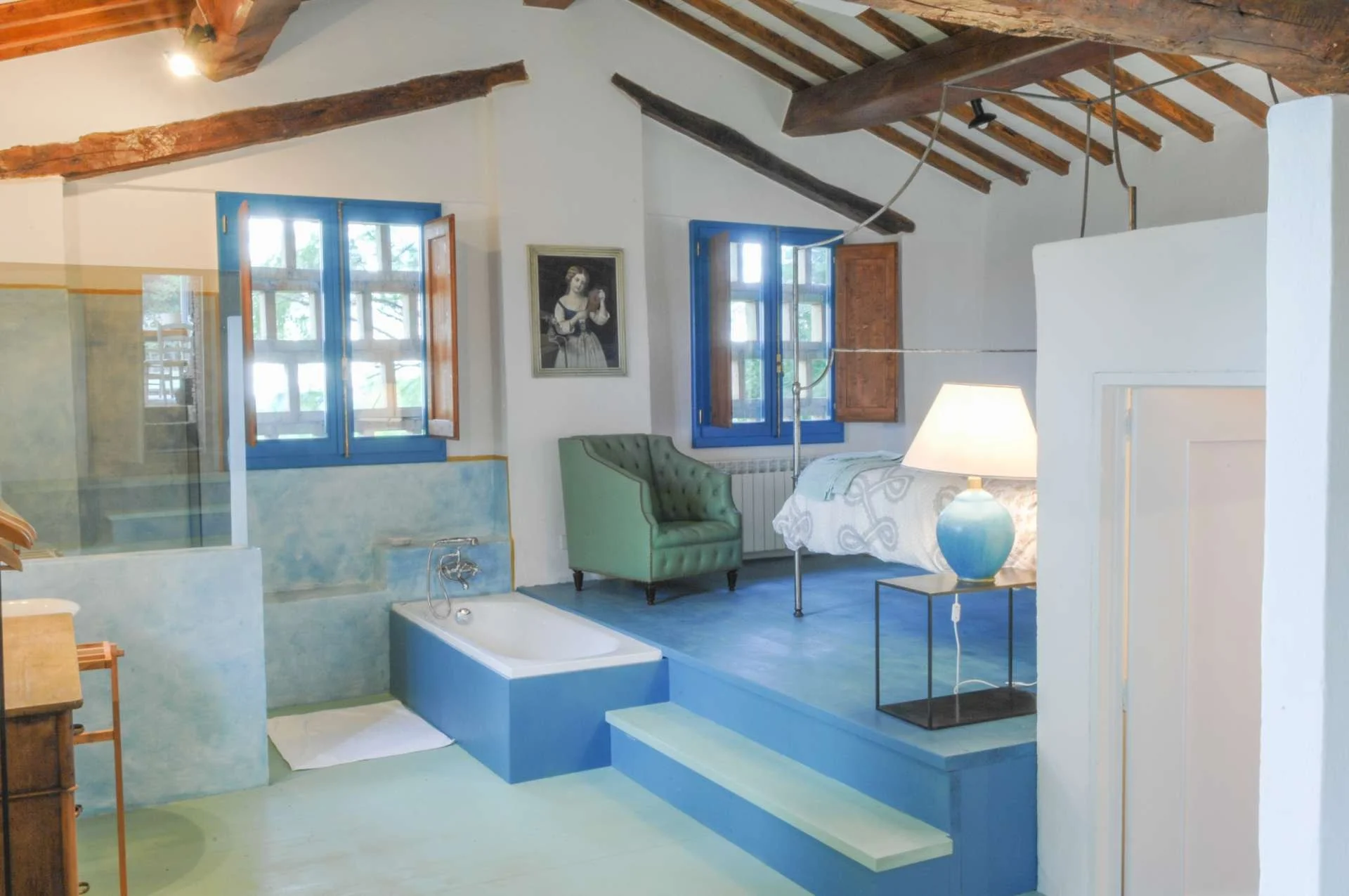 Interior of a rustic-style room with vaulted ceiling and exposed wooden beams. Features include blue window frames, a green tufted armchair, a white bed with a patterned cover, a bedside table with a lamp, and a framed artwork on the wall.