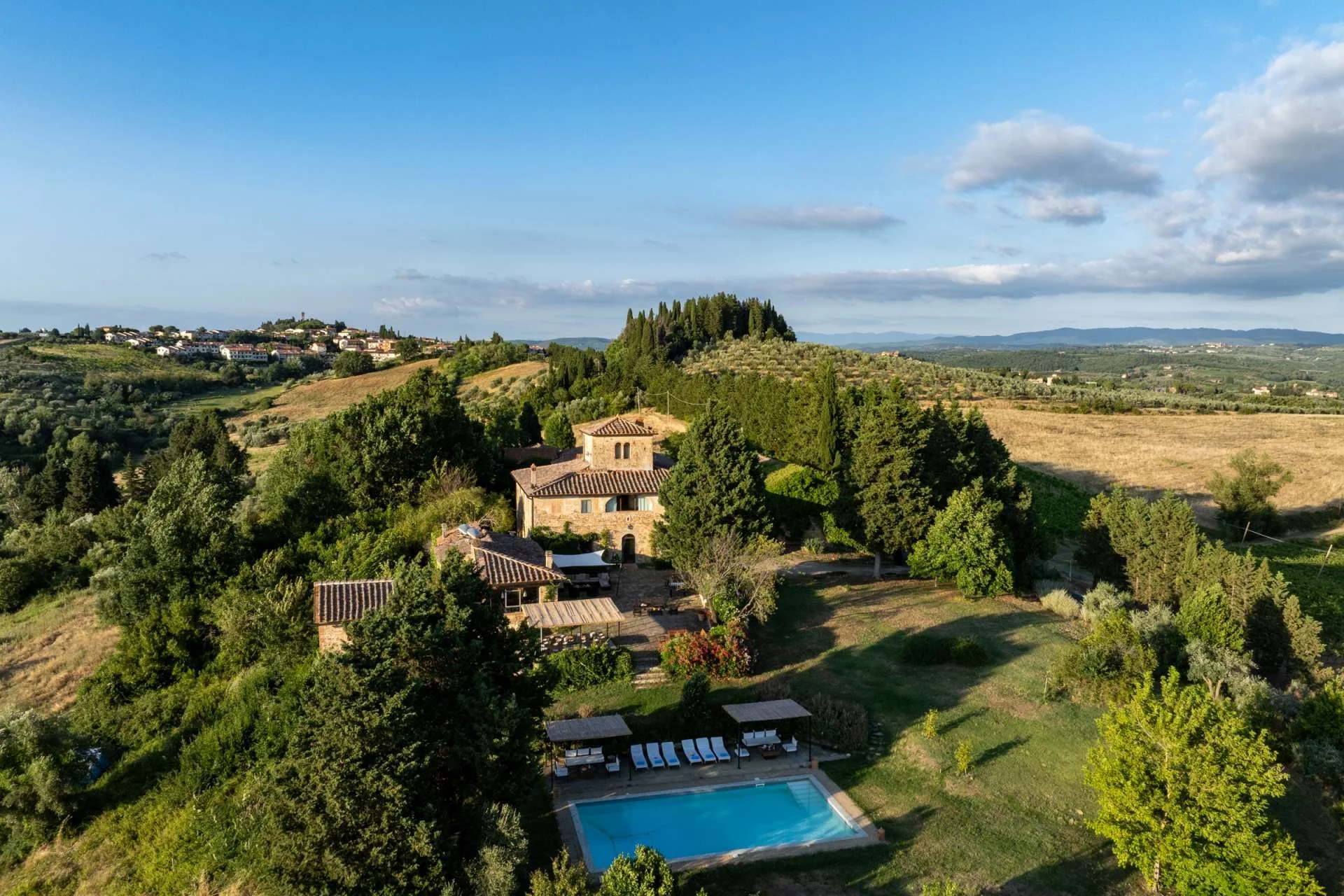 Scenic view of a countryside landscape with rolling hills, trees, a swimming pool with lounge chairs, and a stone house in the foreground, under a partly cloudy sky.