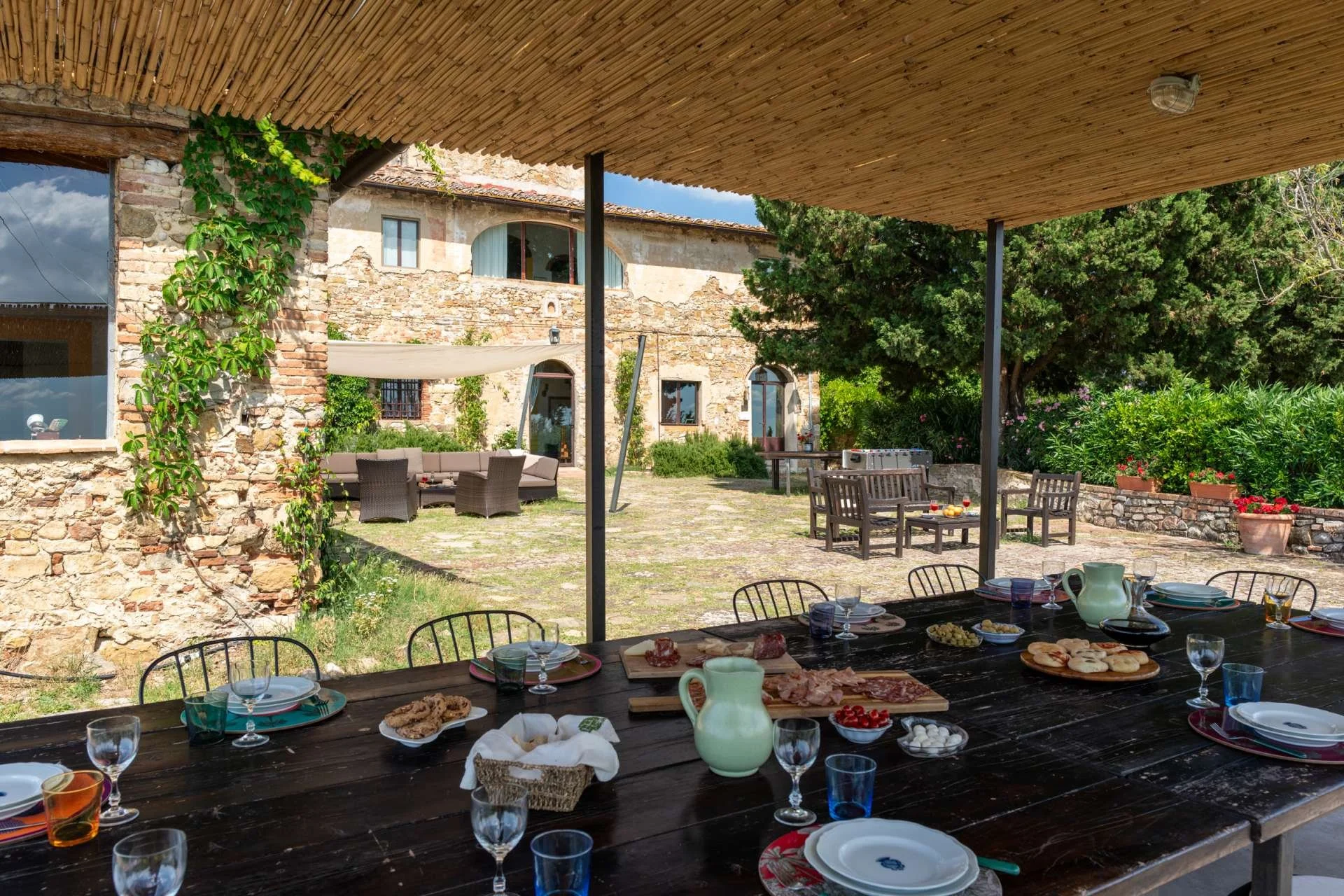 Outdoor patio dining area with a table set with plates, glasses, and food, overlooking a garden with an old stone building in the background.