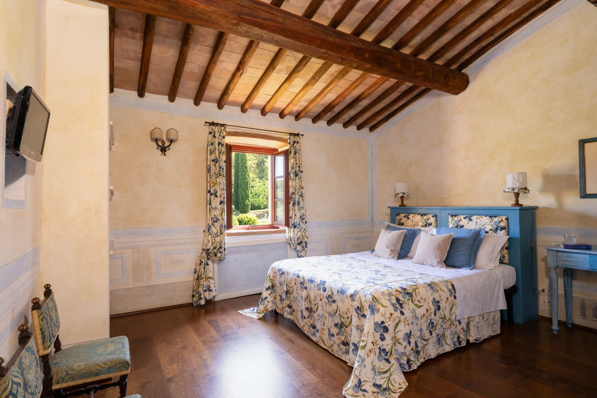A cozy bedroom with a high, wooden-beamed ceiling, a window with floral curtains, and a blue headboard with matching pillows, a nightstand with lamps, and a small chair with floral upholstery.