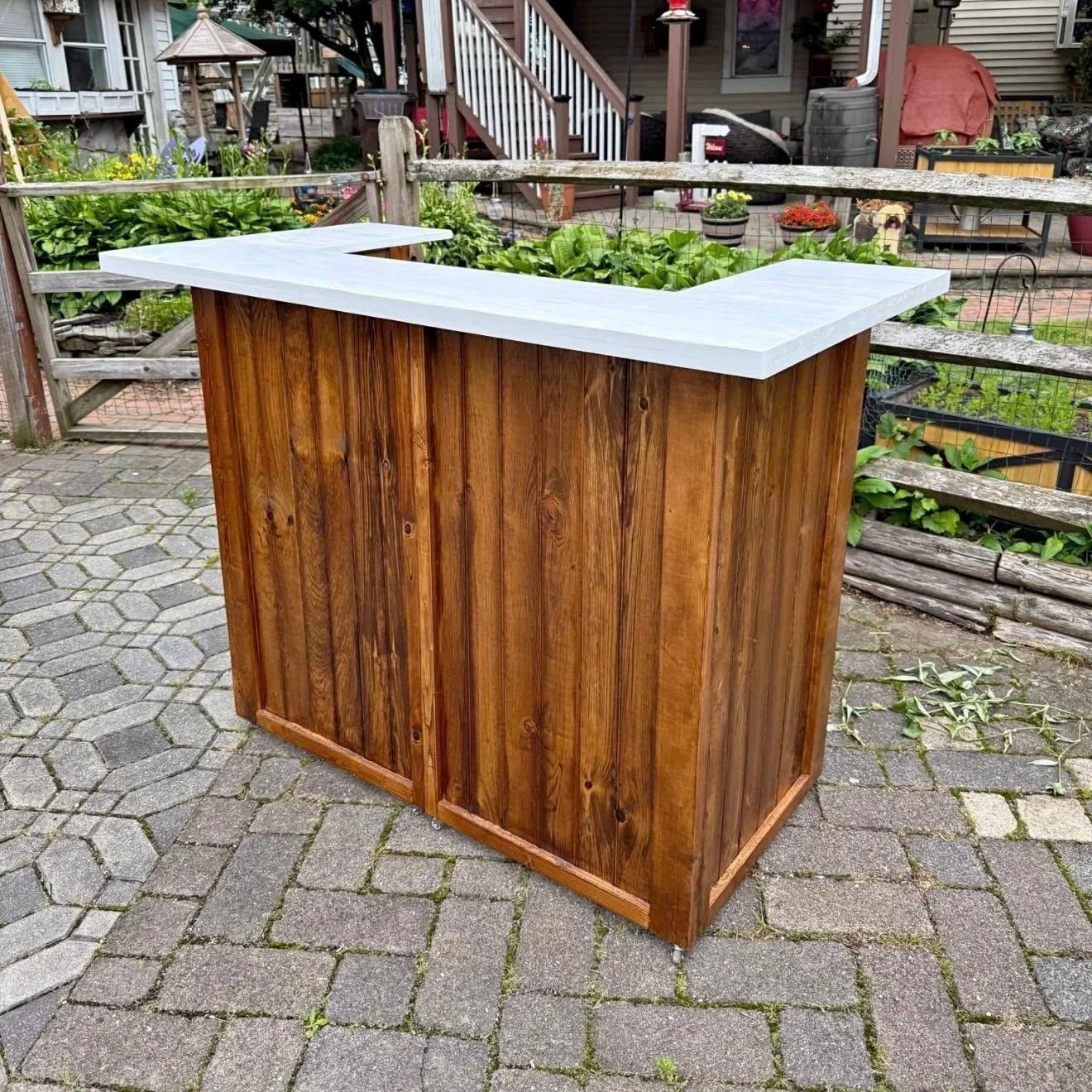 A wooden outdoor bar with a white countertop, set on a stone patio in a backyard garden.