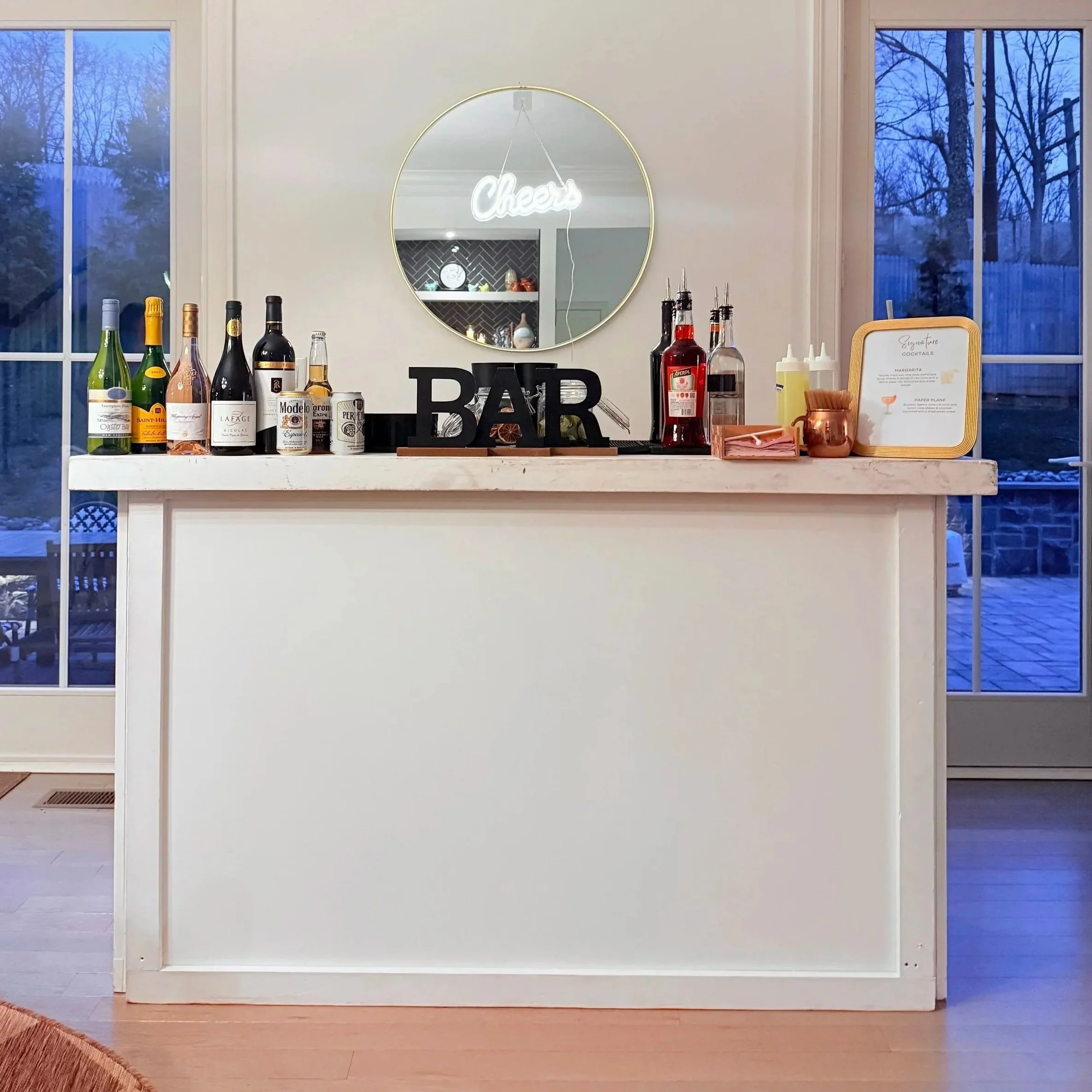 Indoor bar setup with various alcohol bottles, condiments, a mirror with a neon 'cheers' sign, and a sign labeled 'BAR' on a white counter. Large windows reveal an outdoor patio and trees.
