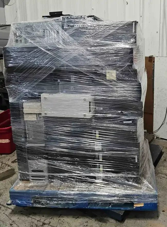 A wooden pallet stacked high with discarded desktop computers and servers, wrapped in clear plastic for secure transport and recycling.