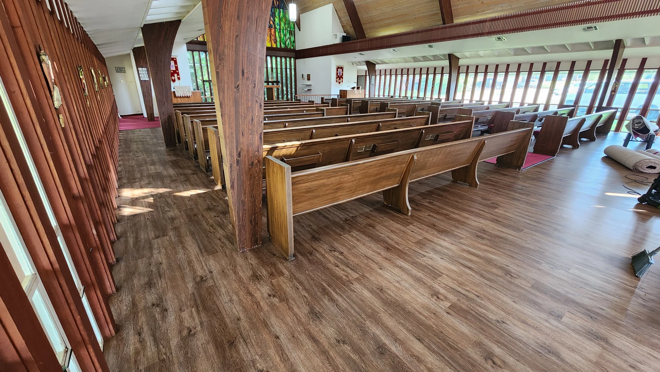 Interior of a church with wooden pews, a stained glass window, and sunlight streaming through large windows along the side.
