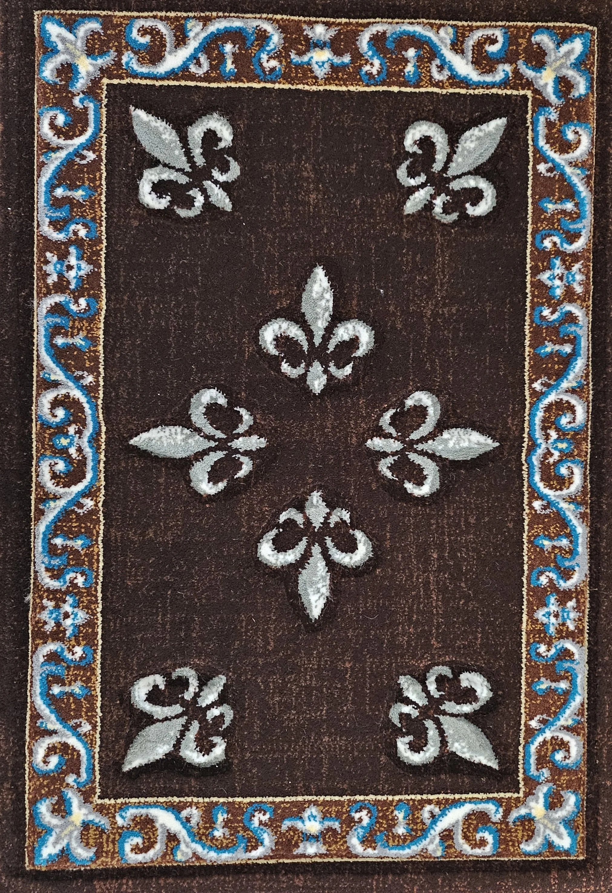 A brown area rug with a decorative border in orange, blue, and white. The border features intricate scrollwork and floral patterns. The main field of the rug displays a pattern of white fleur-de-lis motifs evenly spaced in a diamond arrangement.