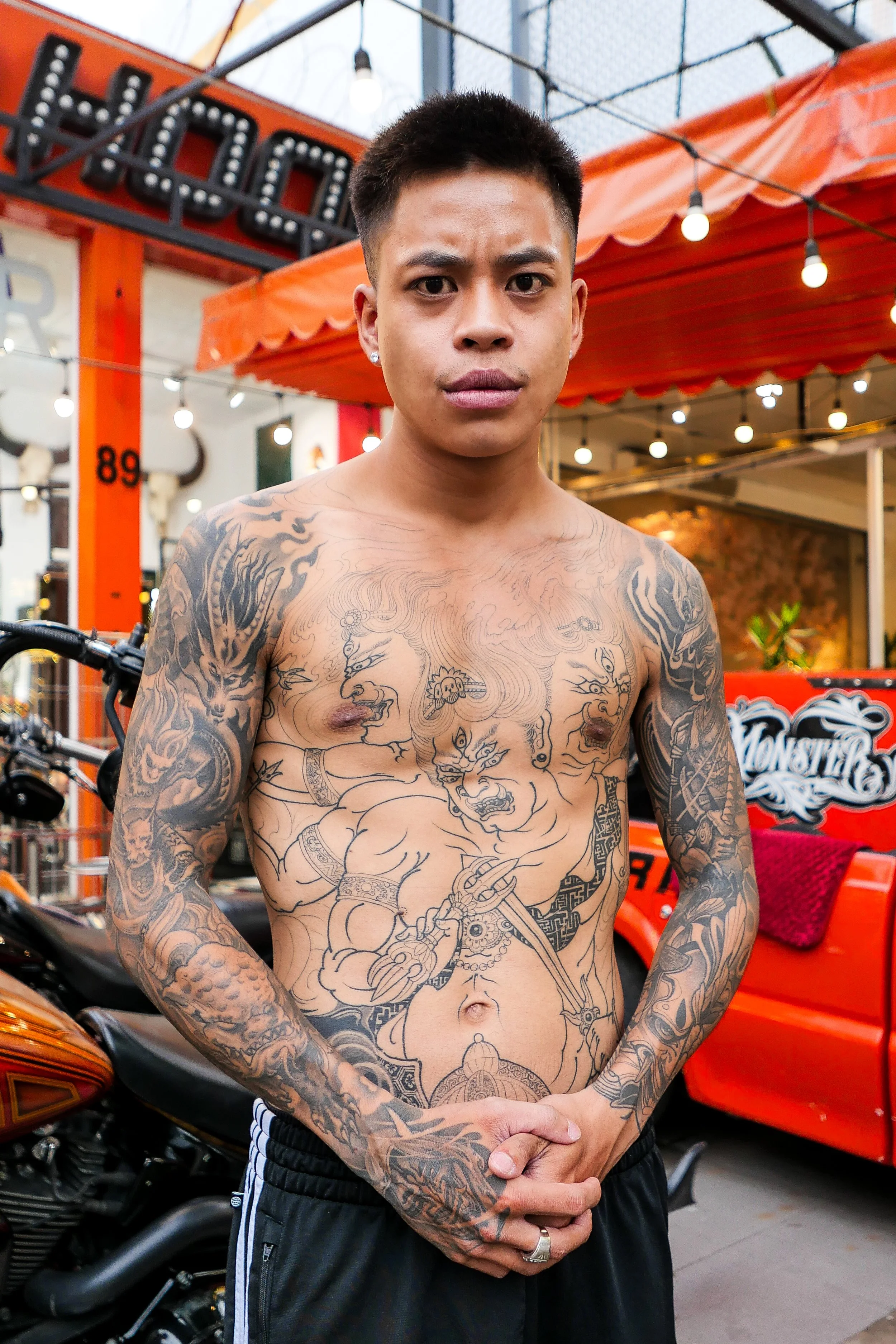A shirtless man with tattoos on his chest and arms, standing in front of orange and black-themed decor, a motorcycle, and a red cart with decoration that says 'Monster'.