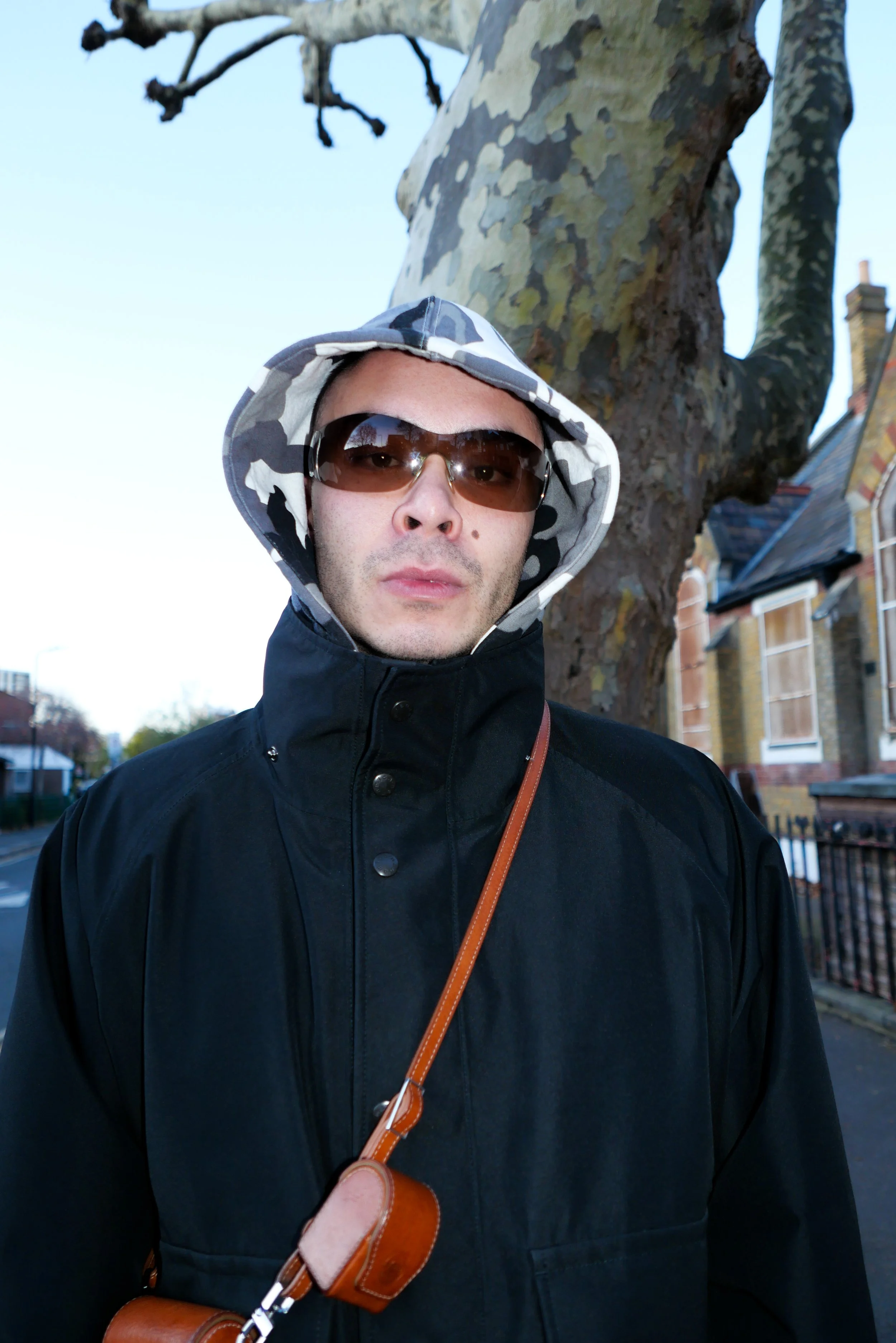 A man wearing sunglasses, a black coat, and a camo hood stands outdoors in front of a large tree with a textured bark. Residential houses with brick and siding are visible in the background.