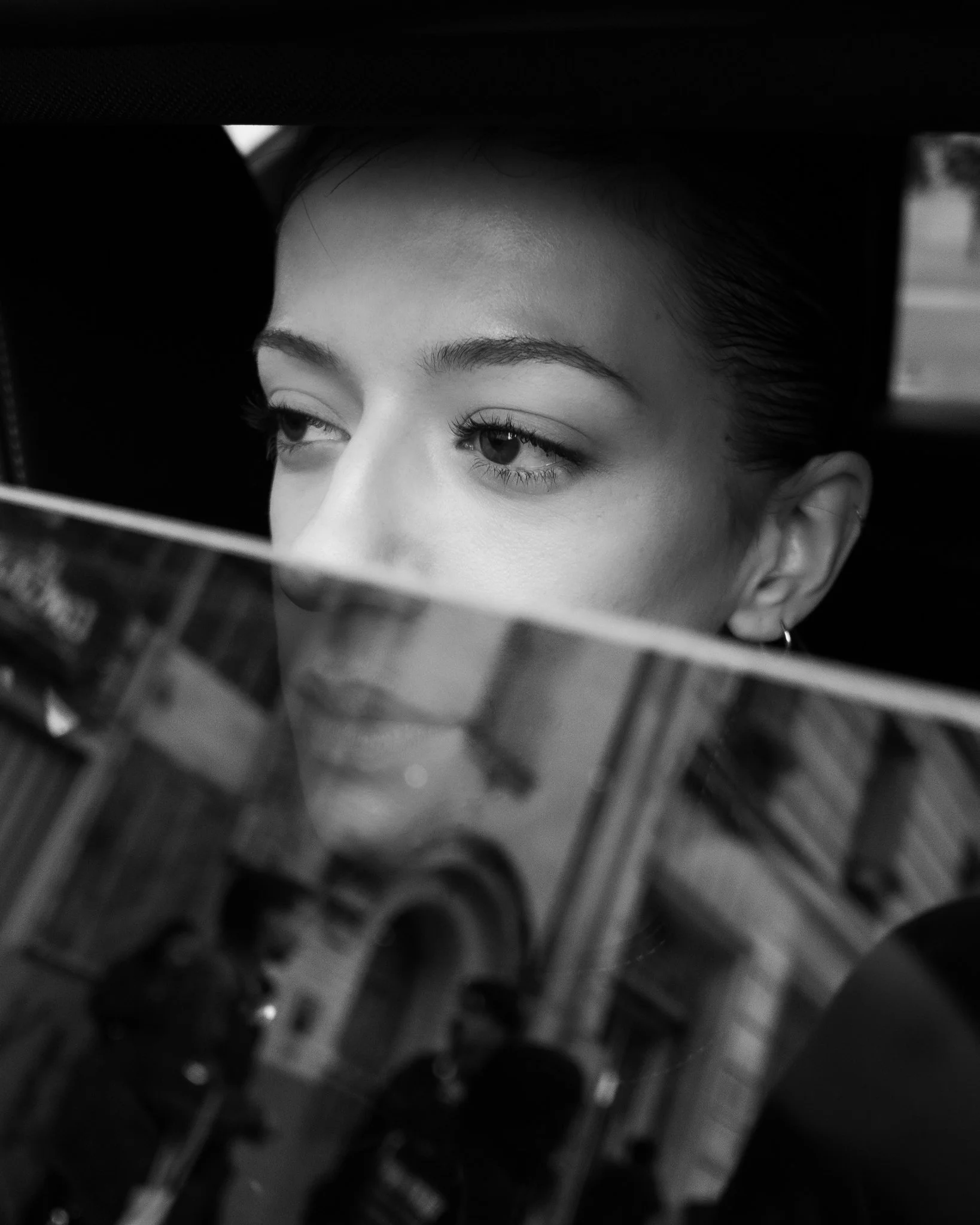 Close-up of a woman's face with makeup, looking to the side, partially obscured by a reflective surface.