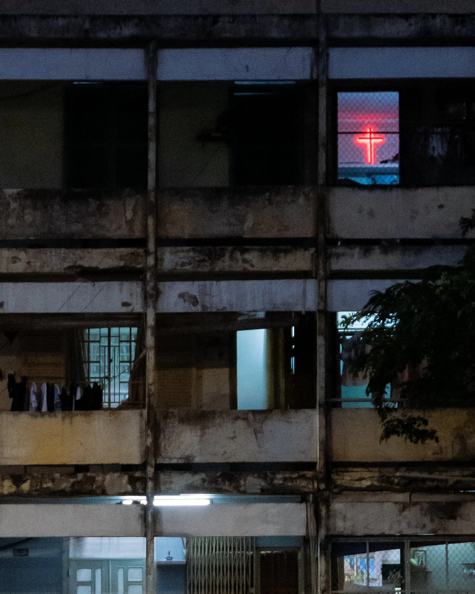 Nighttime view of an apartment building with a glowing red neon cross in the window at the top.