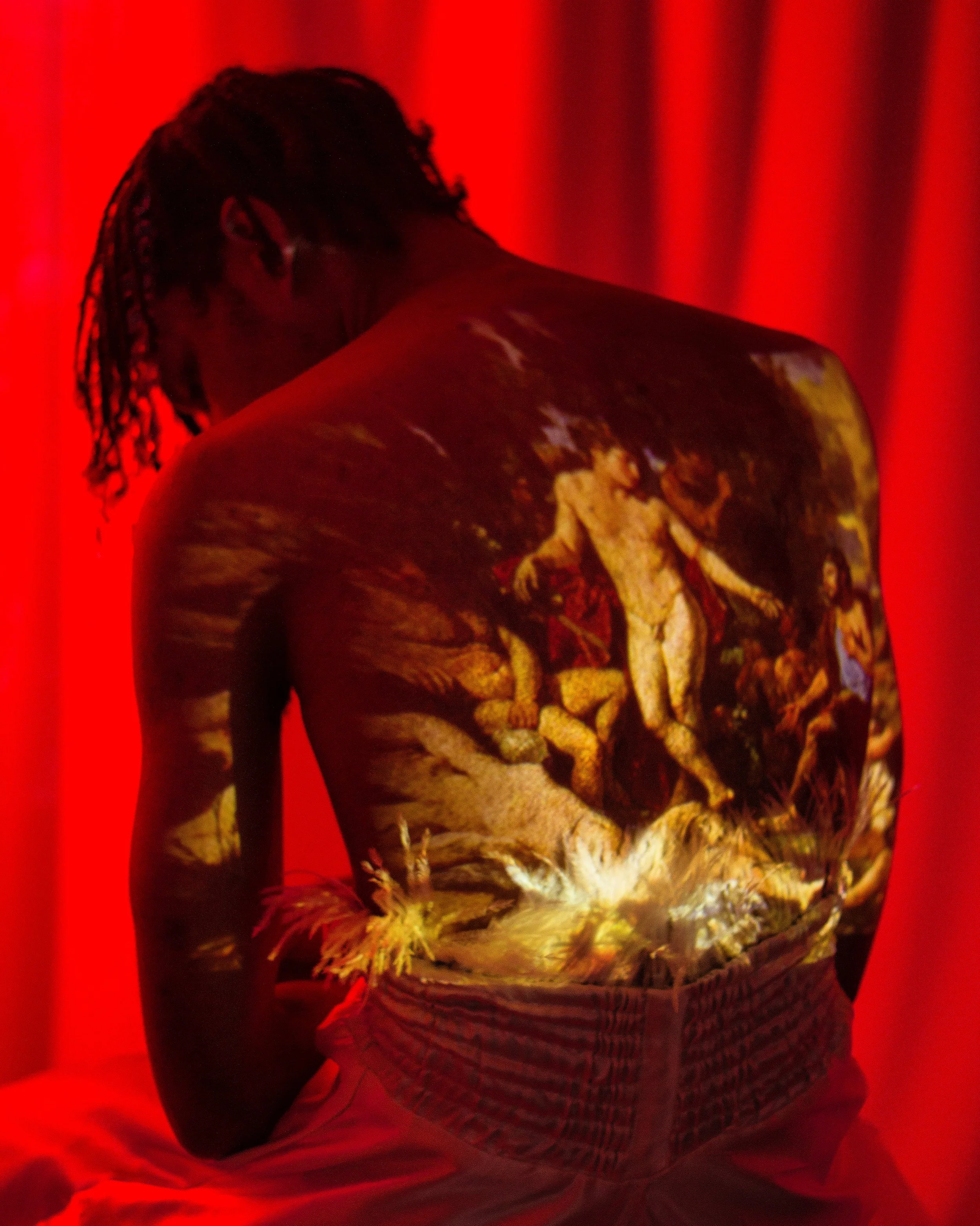 A person with dreadlocks facing downward has a painting of Michelangelo's "The Creation of Adam" projected onto their back, set against a red background.