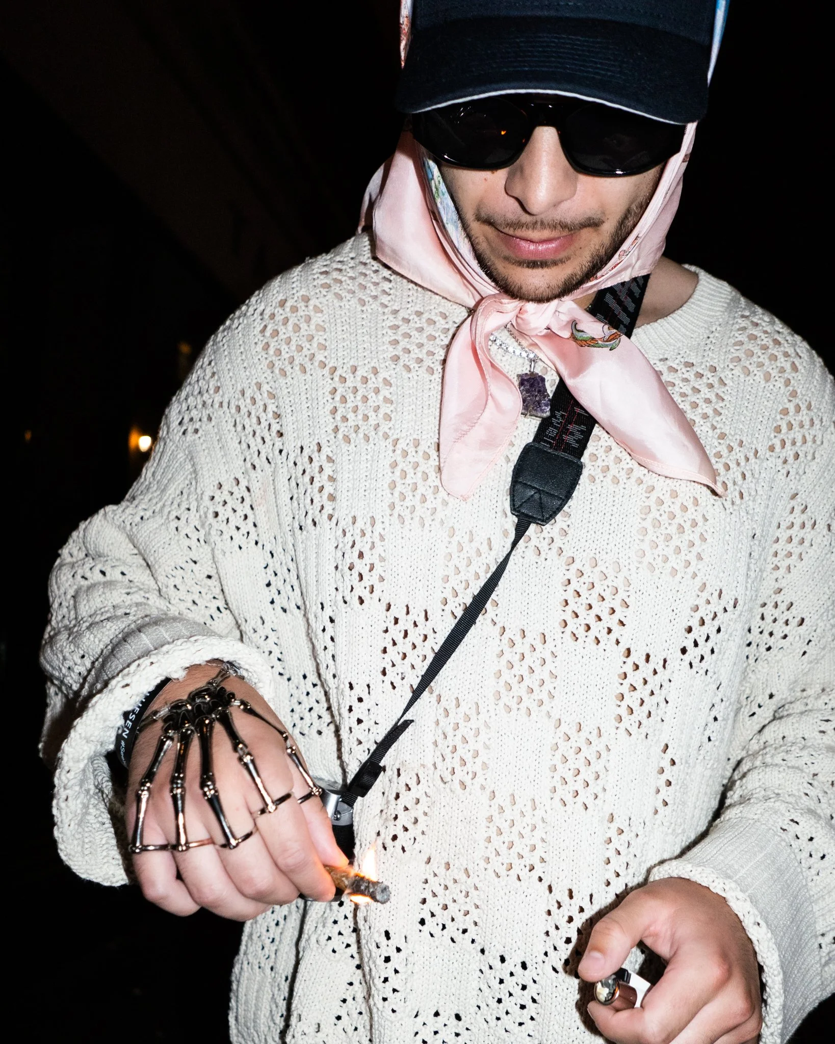 A man wearing sunglasses, a pink scarf, and a white crocheted sweater, holding a lit cigarette and a lighter, with some jewelry including rings and a bracelet.