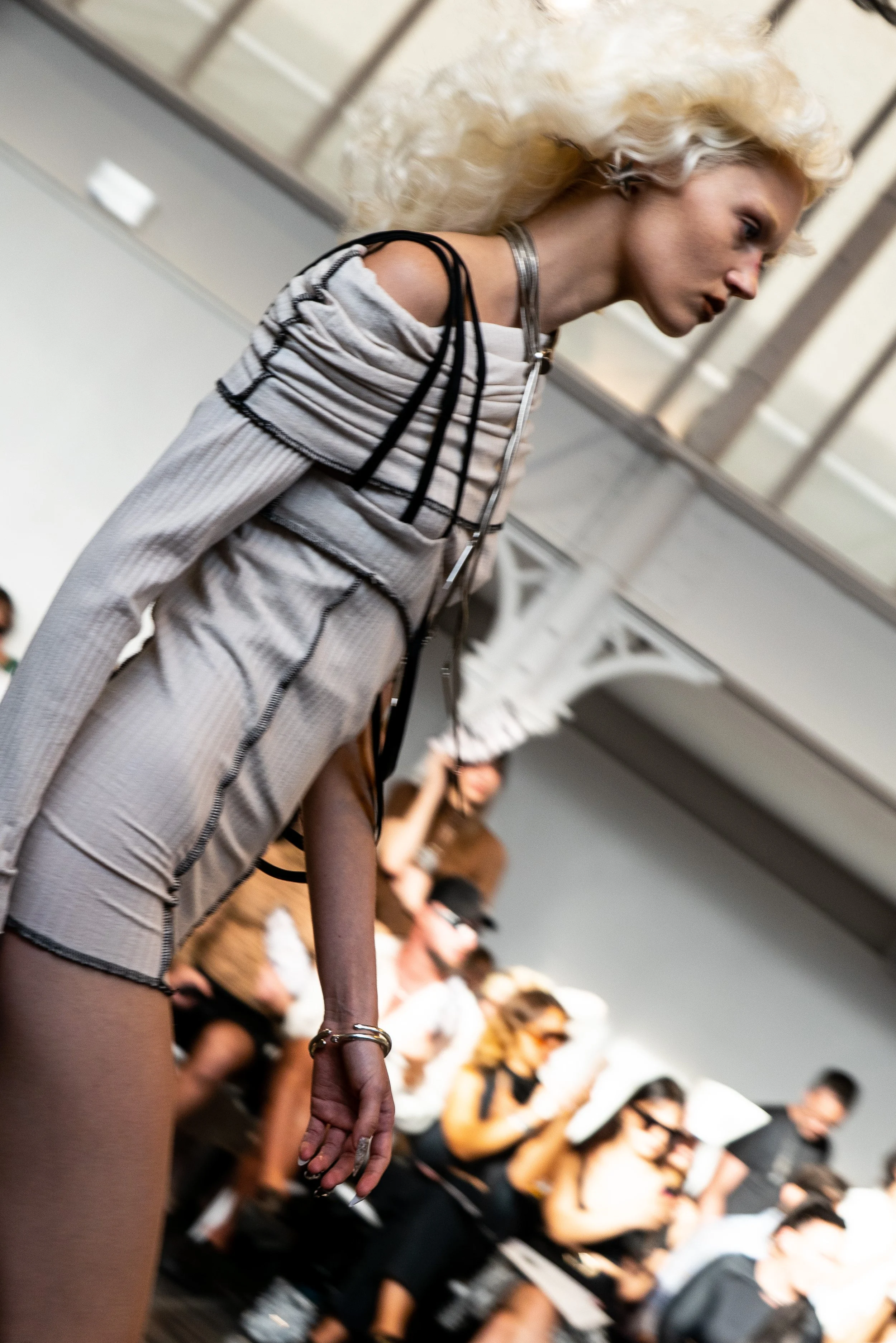 Fashion model with platinum blonde hair walking on runway during a fashion show, wearing a grey and black striped outfit with cut-out shoulders, in front of an audience with some people taking photos.
