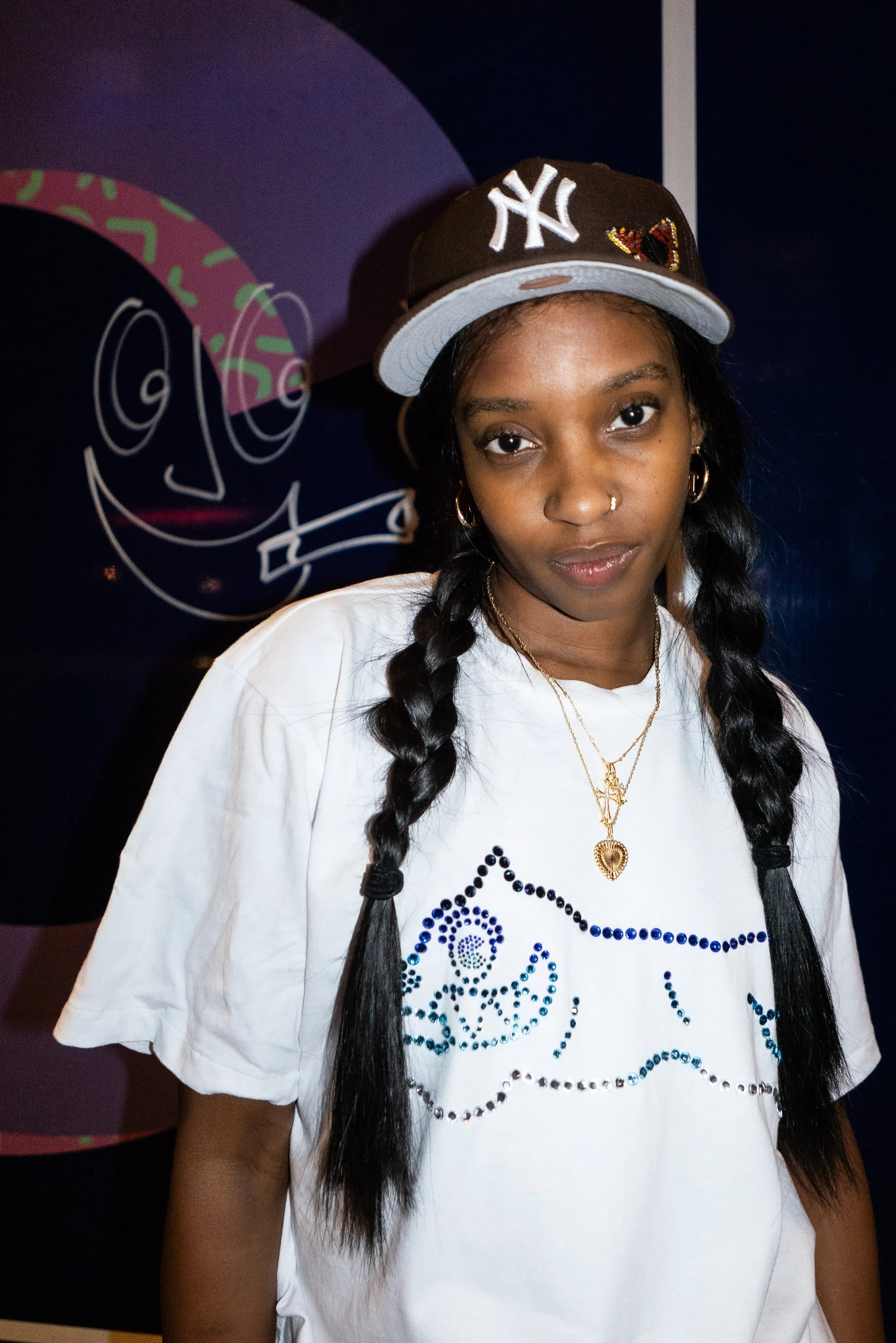 A young woman with braided hair, wearing a New York Yankees cap and a white T-shirt with rhinestone designs, stands in front of a colorful wall with a face drawing.
