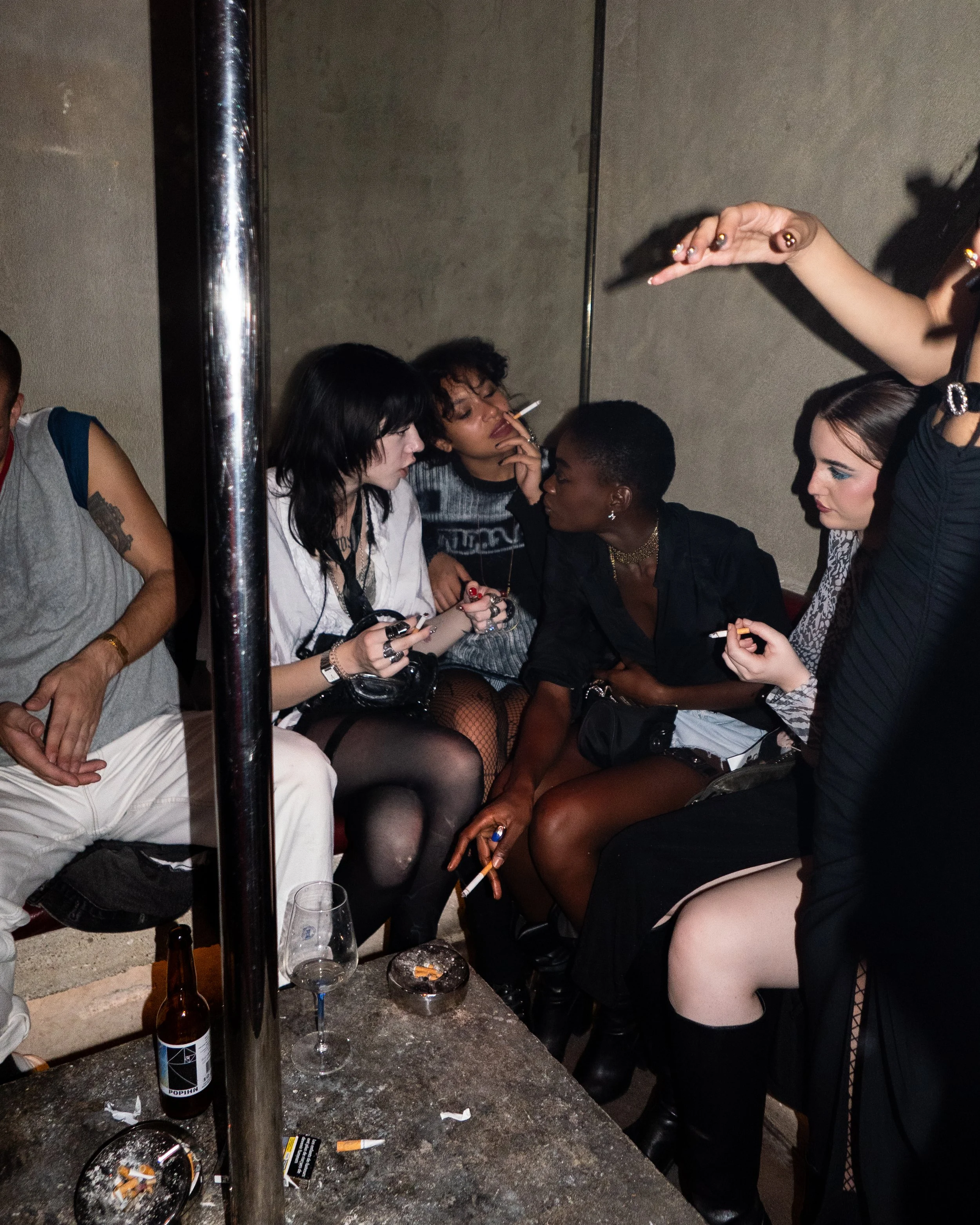 Group of young people sitting on a couch in a dimly lit room, smoking cigarettes and engaging in conversation.