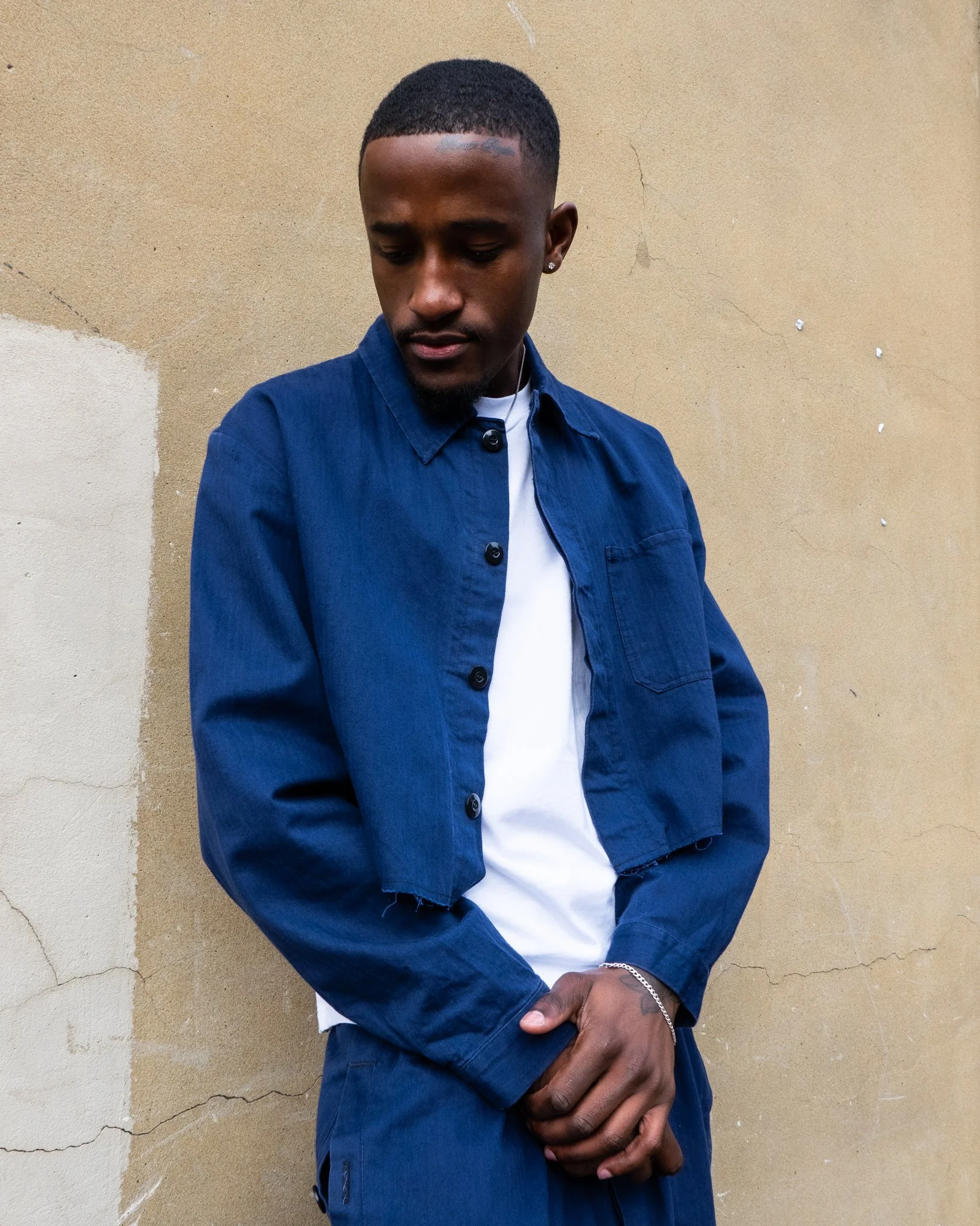 A young man with short hair and earrings, wearing a blue denim jacket over a white shirt, standing against a beige cracked wall with his hands clasped in front of him, looking down.