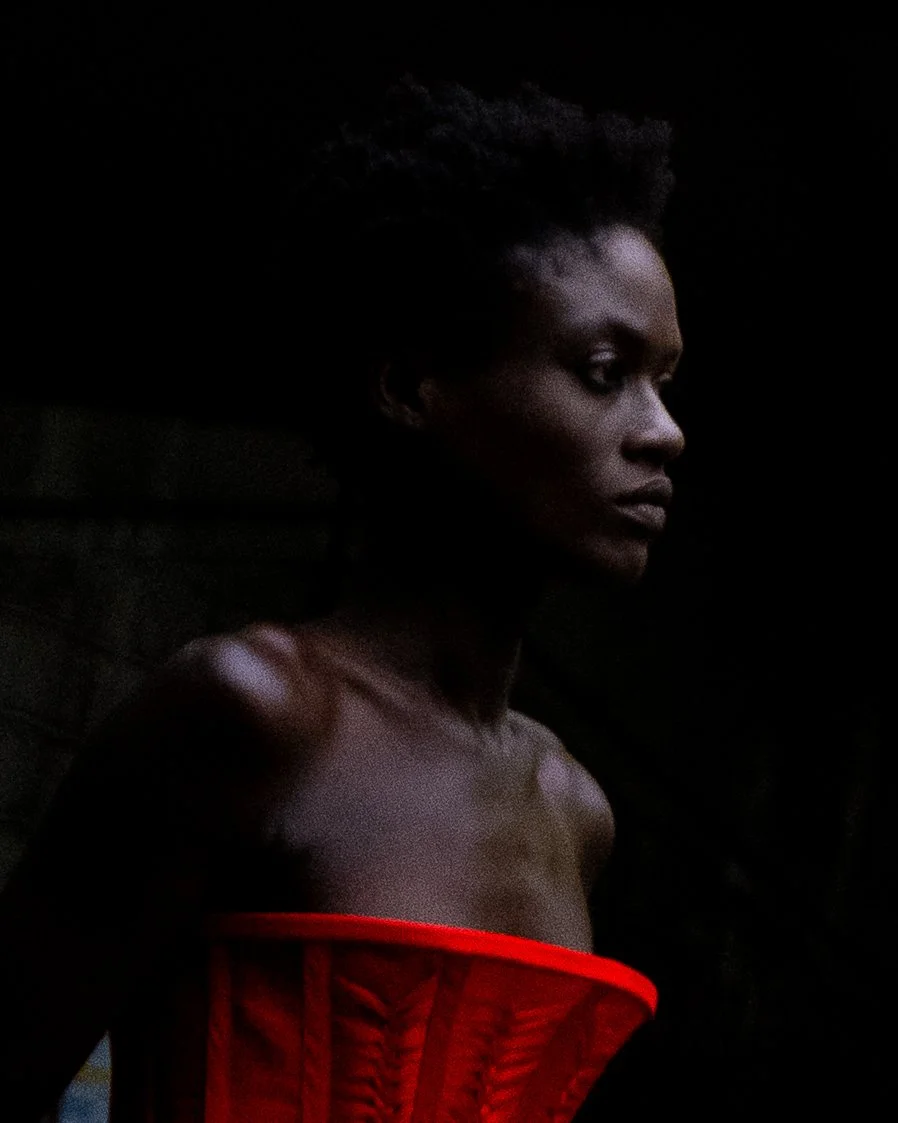 A woman with short curly hair and dark skin wearing a red strapless top, standing against a dark background, looking to the right with a serious expression.