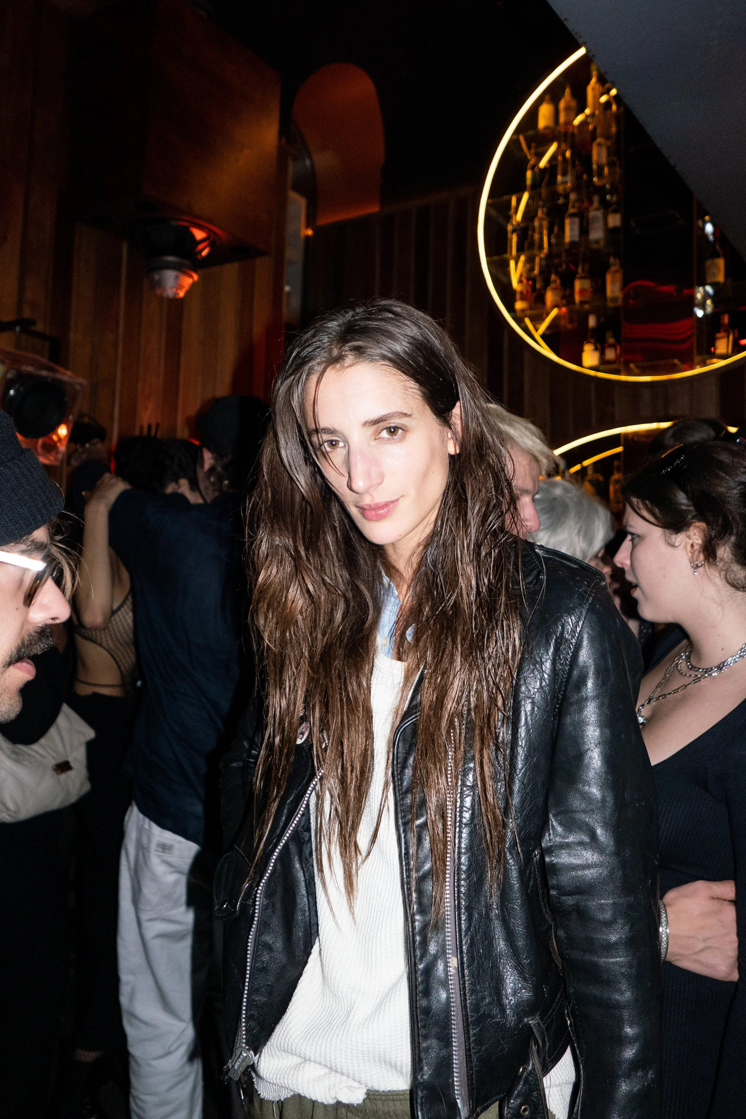 A woman with long brown hair wearing a black leather jacket looks at the camera in a crowded bar or club with wooden walls and circular shelves of liquor bottles in the background.