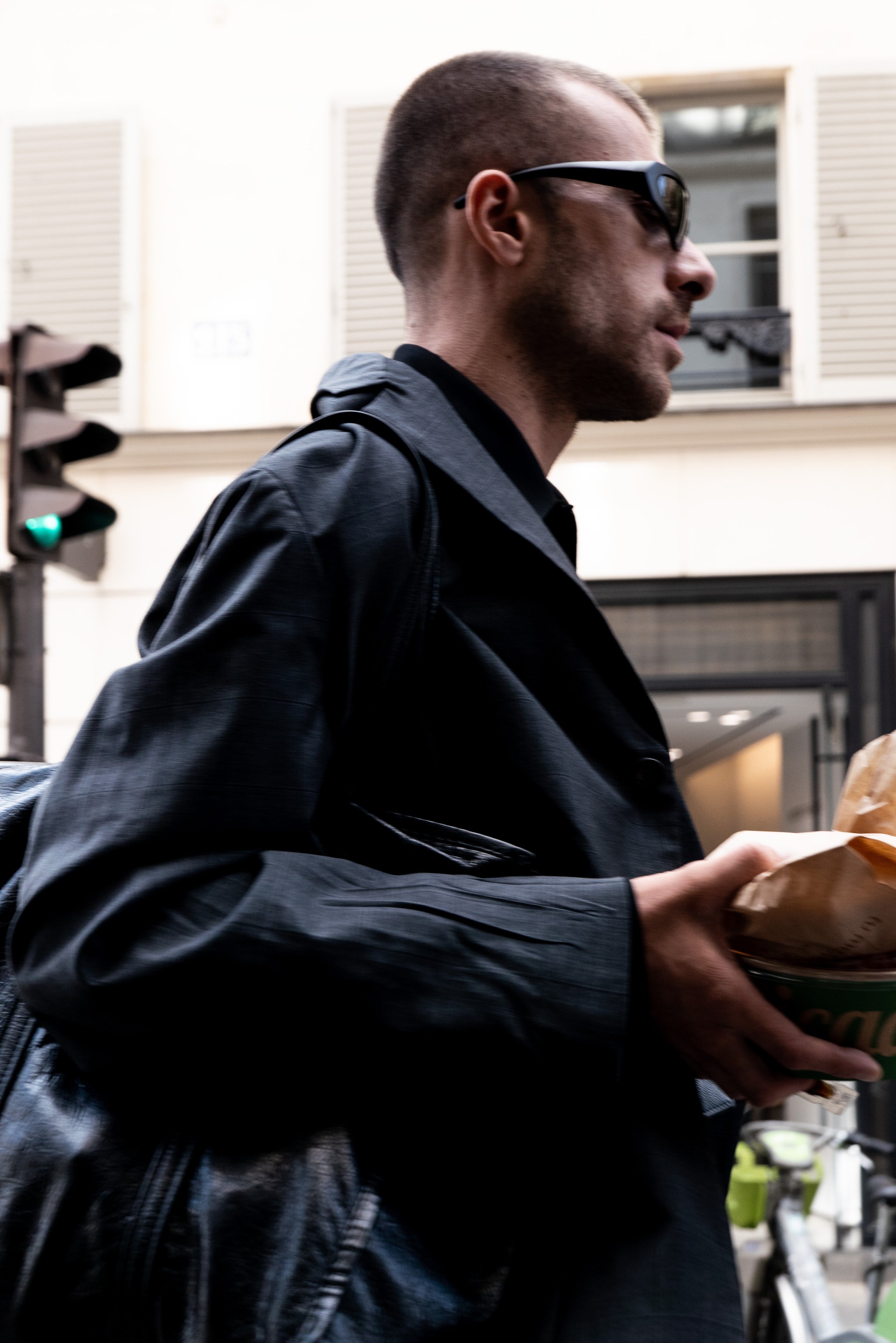 A young man with short hair and sunglasses wearing a black leather jacket holding a takeout container in an urban street scene.