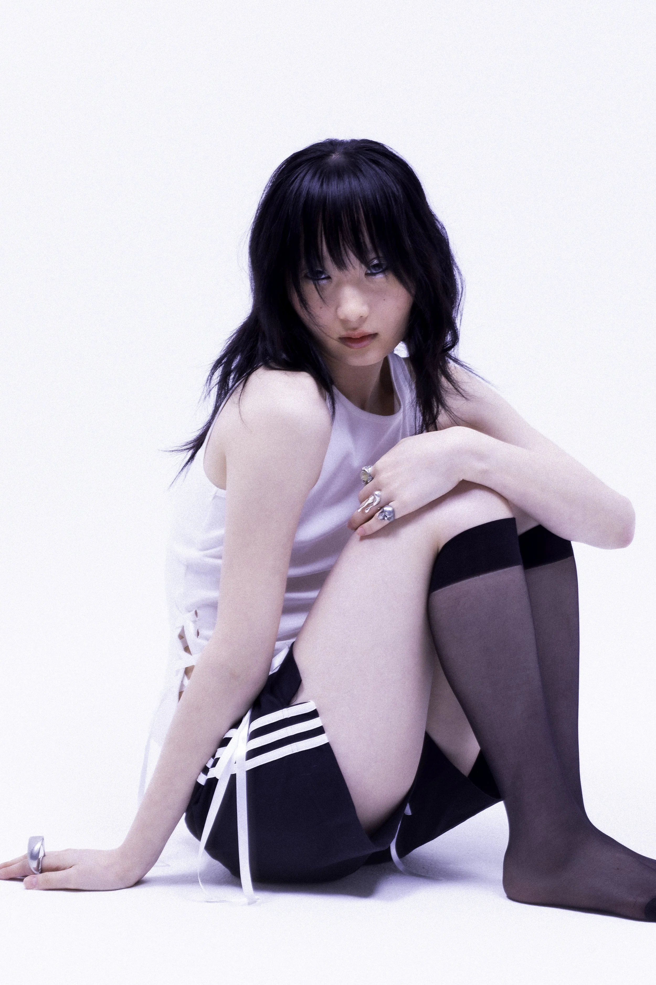 A young woman with black hair and blue eyes sitting on the floor against a plain white background, wearing a white sleeveless top, black and white striped shorts, and black thigh-high stockings.