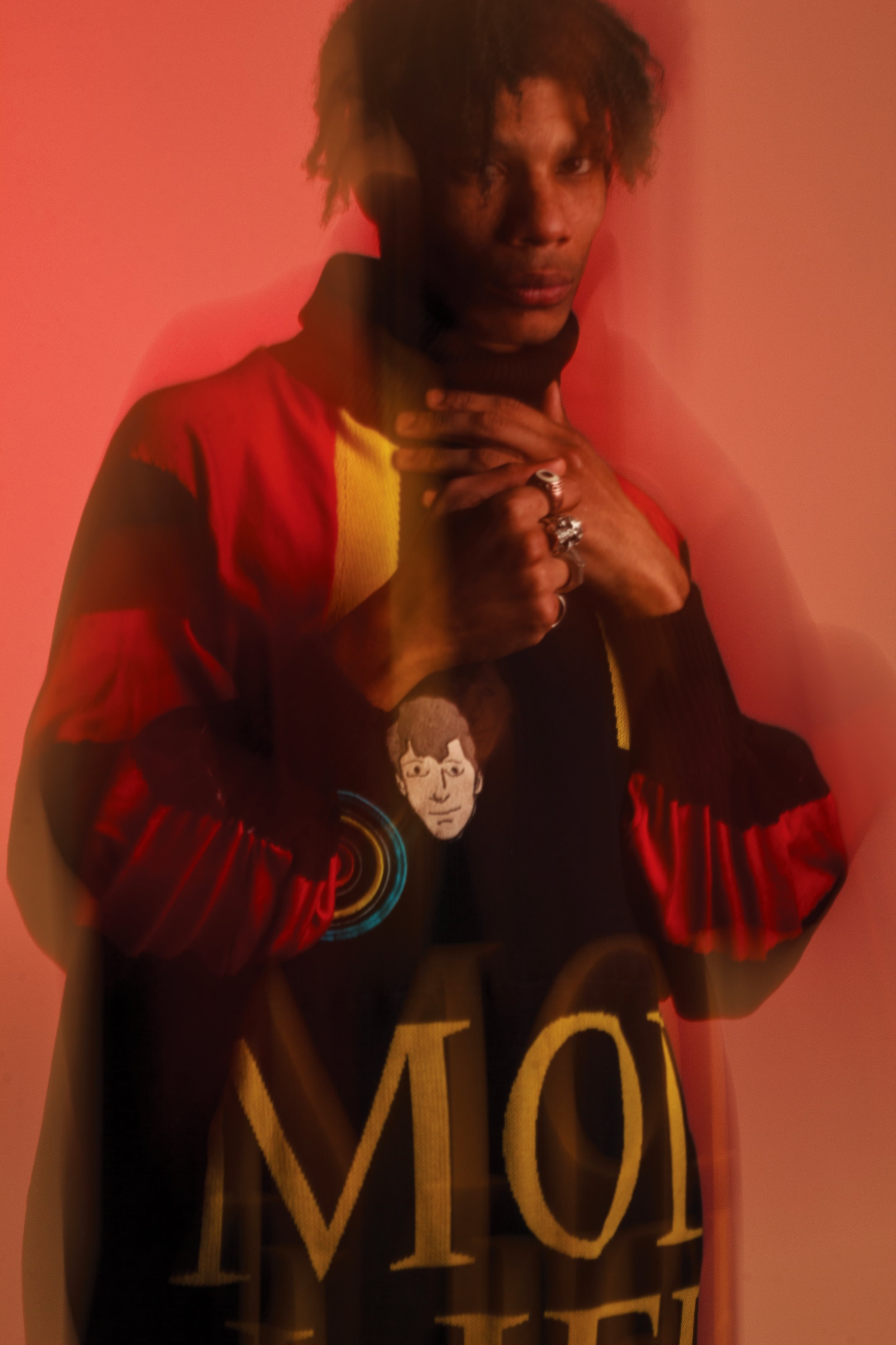 A person standing against a pink background, wearing a colorful jacket with a cartoon face on it, multiple rings, and making a gesture with hands near their neck.