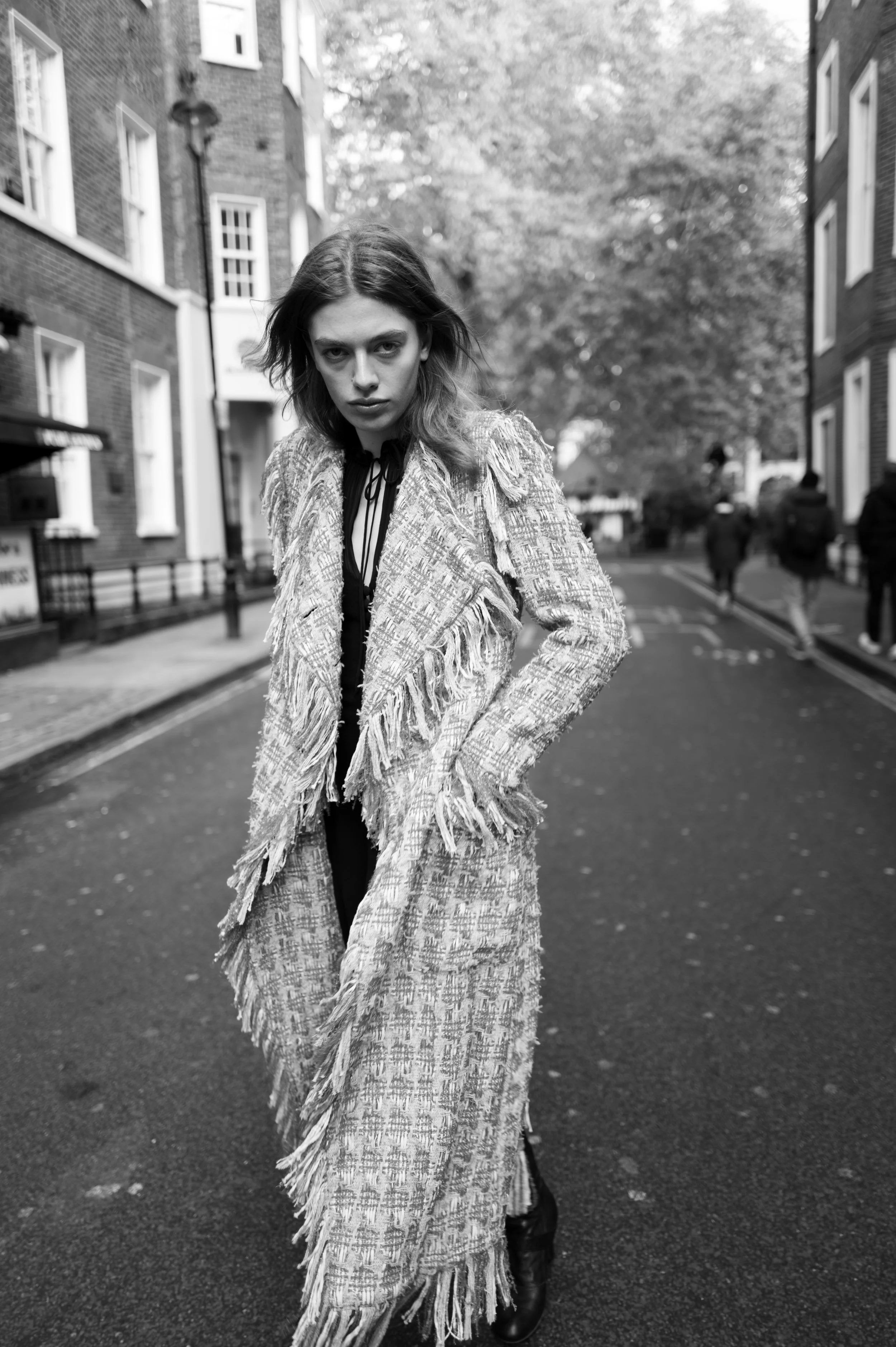 A woman with shoulder-length hair stands on a city street in black-and-white photography. She wears a textured, fringed coat and has a serious expression, with her hands in her coat pockets. In the background, there are buildings, trees, and a few pedestrians walking along the street.