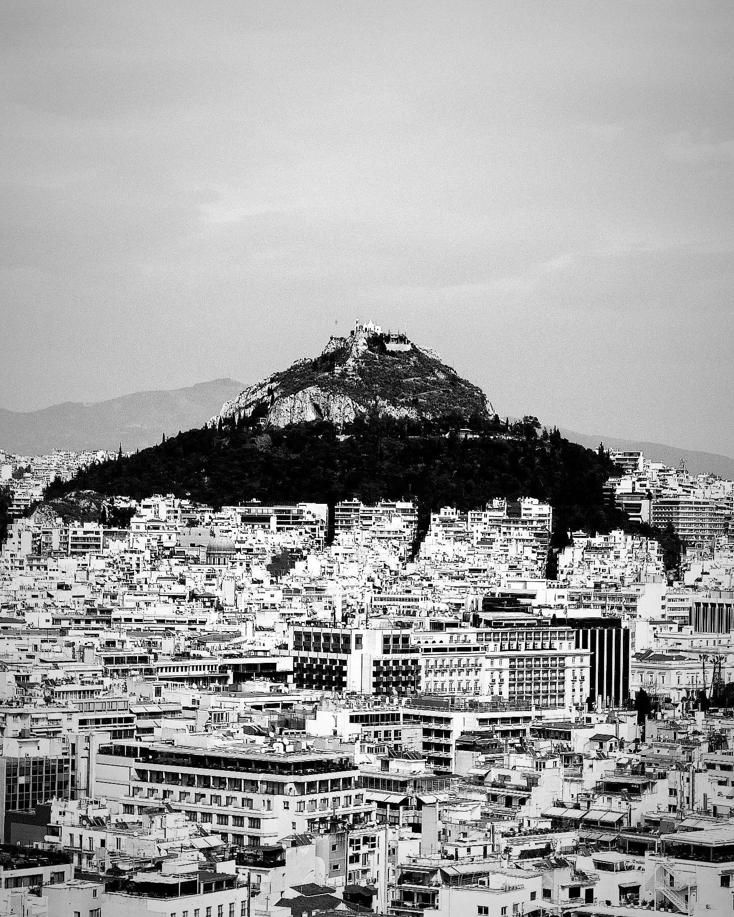 Black and white photo of a city with a hill and a castle on top, showing densely packed buildings and mountains in the background.