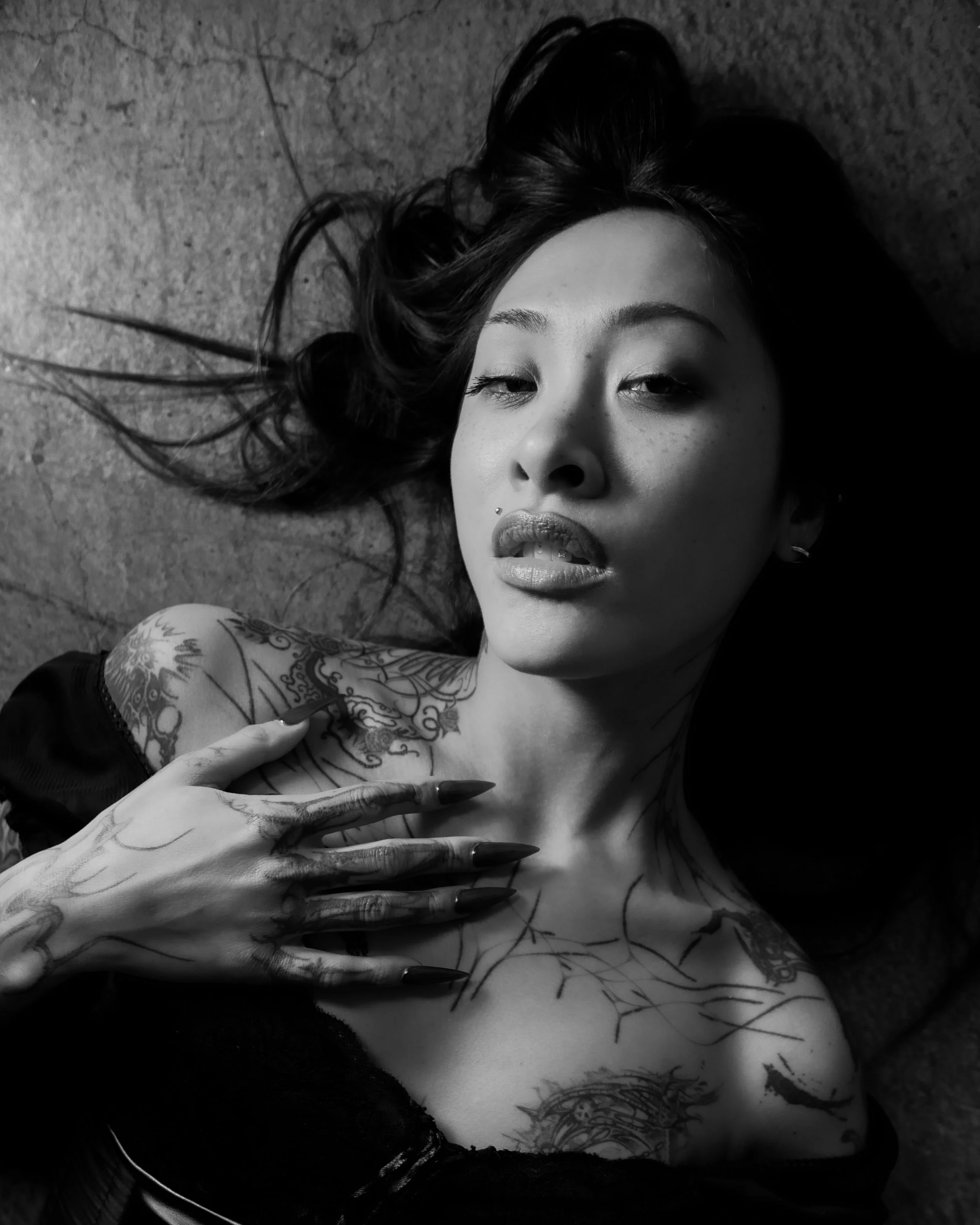 A woman with dark hair lying on the ground, showing tattoos on her arm, chest, and shoulder, with her hand on her chest and her face partially turned towards the camera in black and white.