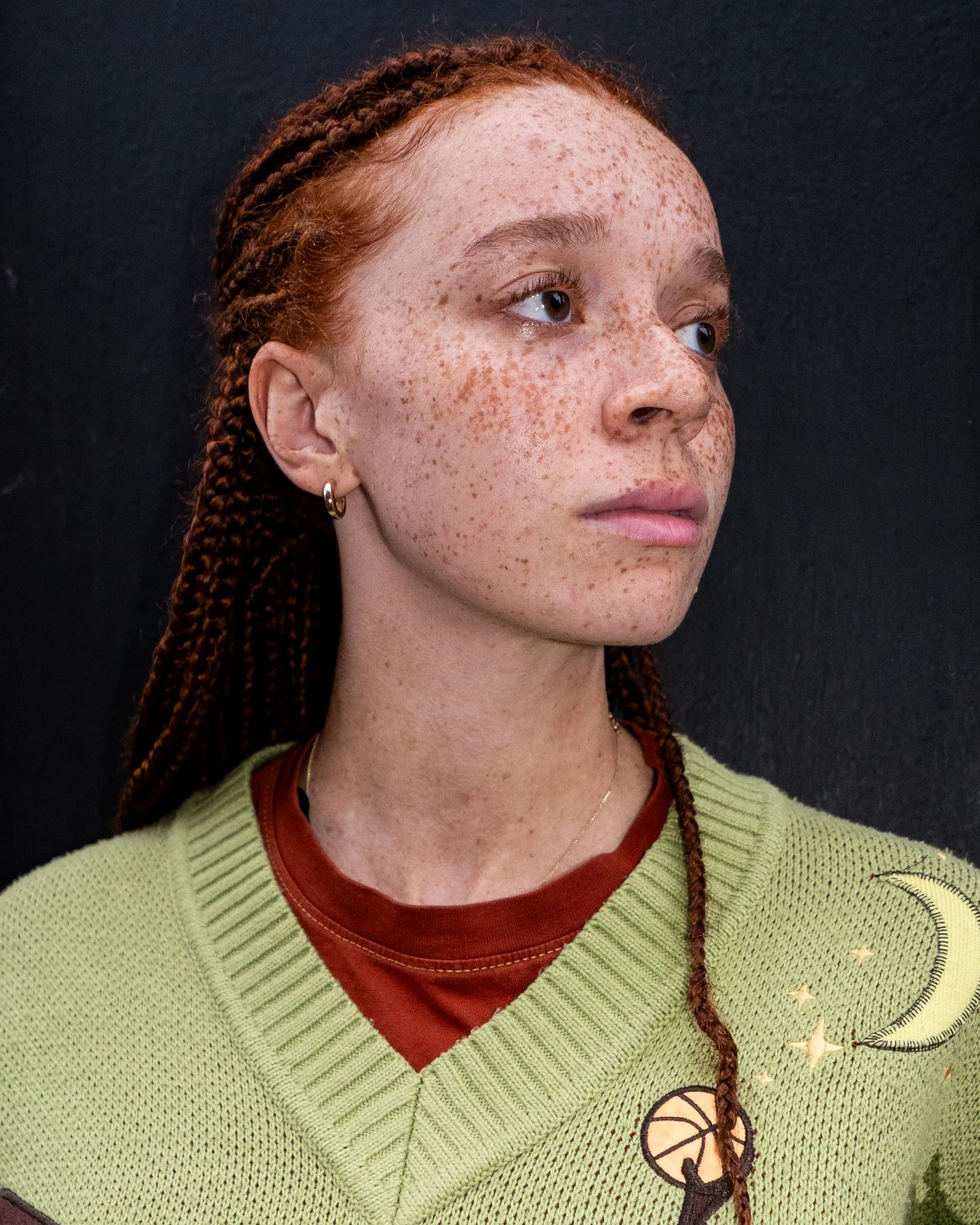 A young woman with red braided hair, light skin, and freckles, wearing a green sweater with celestial patches over a red shirt, standing against a dark background.
