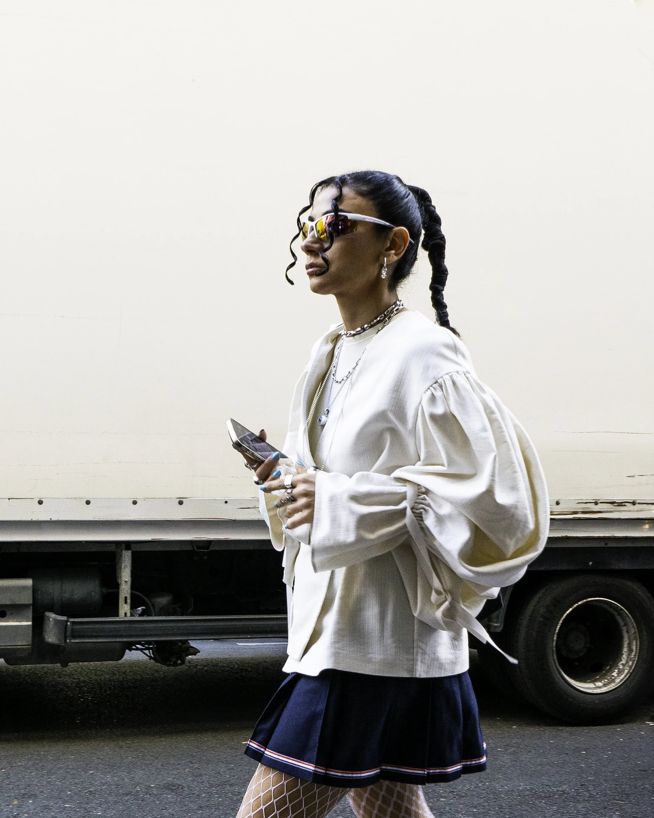 A stylish woman wearing sunglasses, a white jacket, and a black skirt with fishnet tights, holding a smartphone and walking in front of a white truck.
