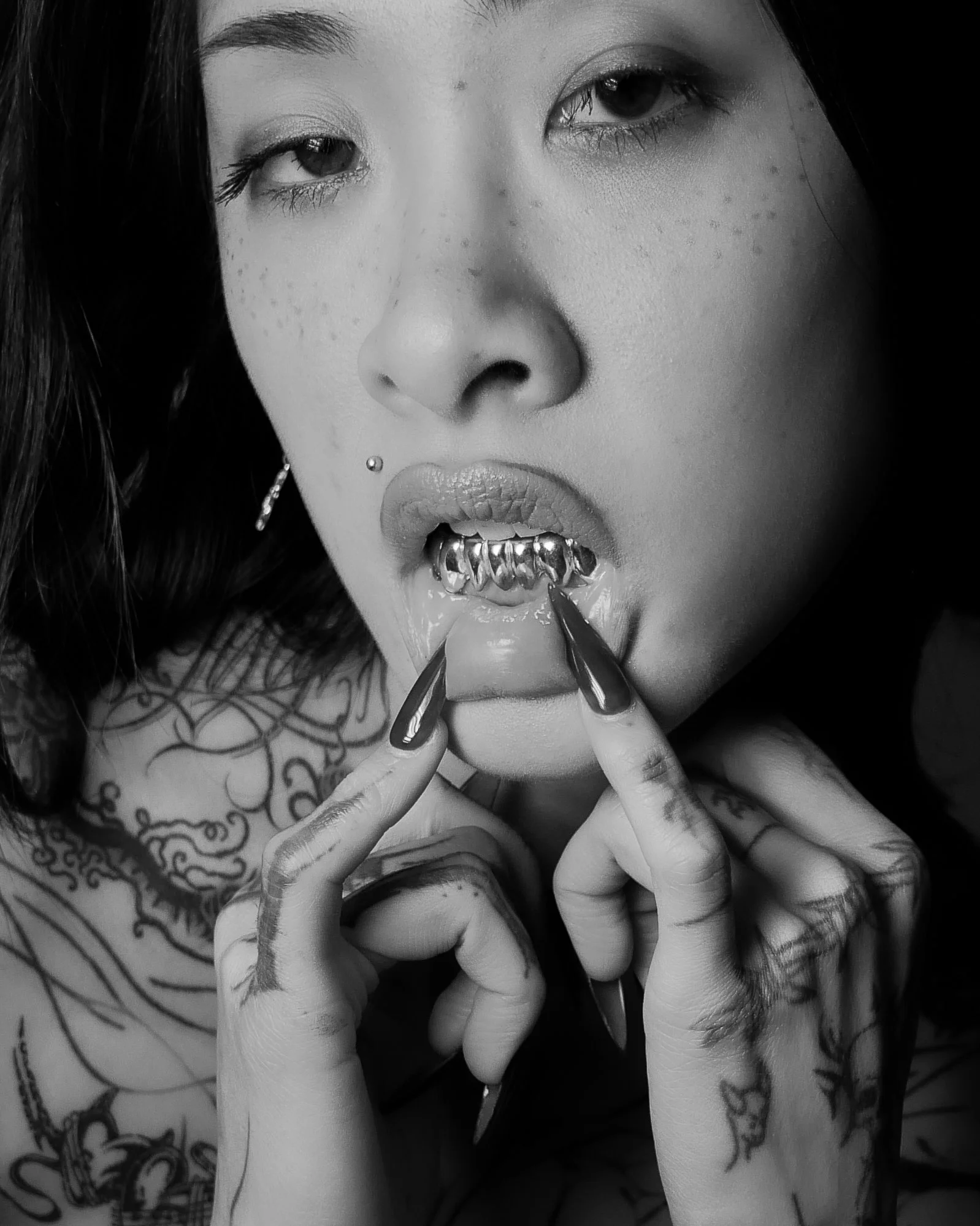Close-up of a woman with tattoos, piercing, dark hair, and dark nail polish, touching her lip with her fingers, showing a chain necklace, piercings, and facial freckles.
