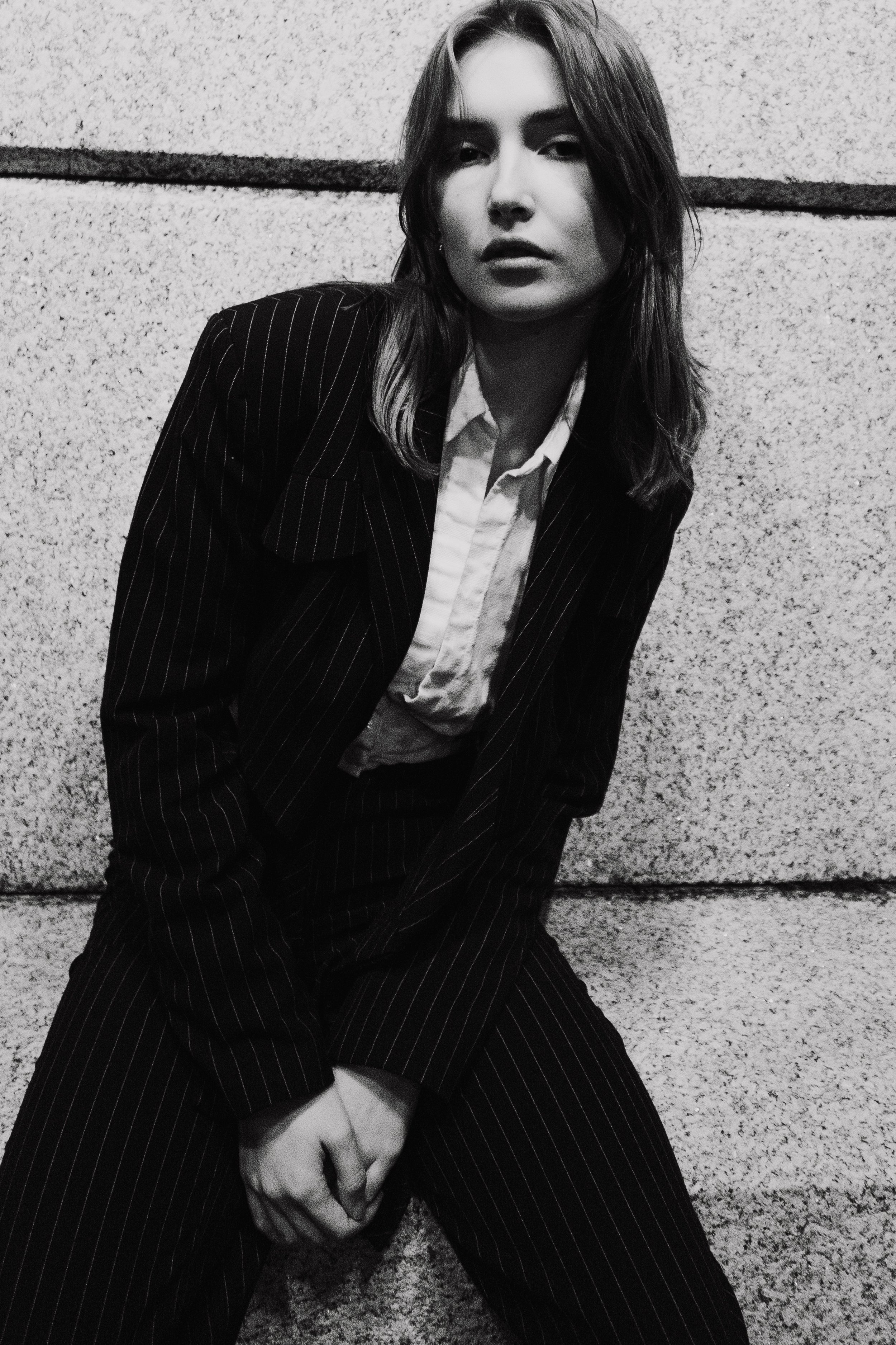 Black and white photo of a woman in a pinstripe suit with disheveled hair, sitting against a textured wall.