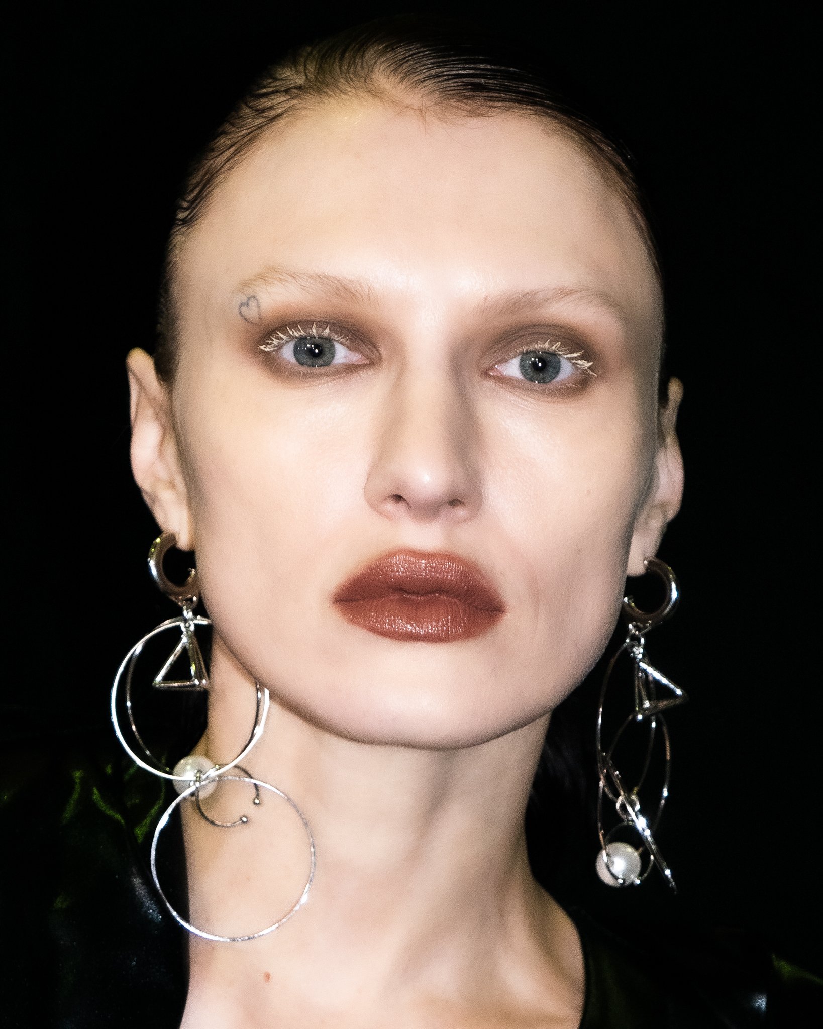 Close-up portrait of a woman with makeup, earrings, and a small heart tattoo near her eyebrow, against a dark background.