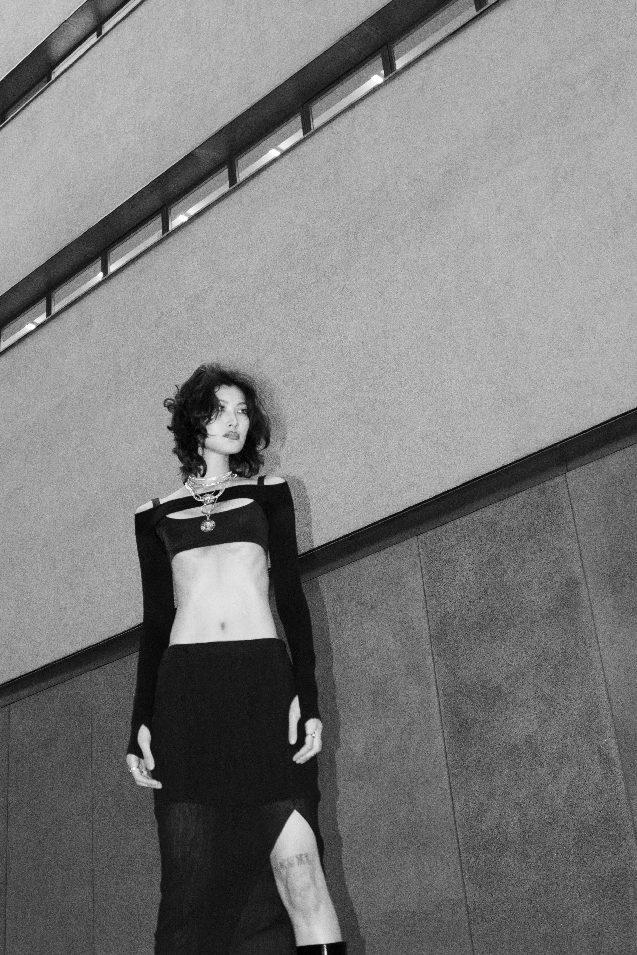 A woman with curly hair standing against a modern building wall, wearing a black cut-out long-sleeve crop top, a black skirt with a high slit, and layered necklaces.