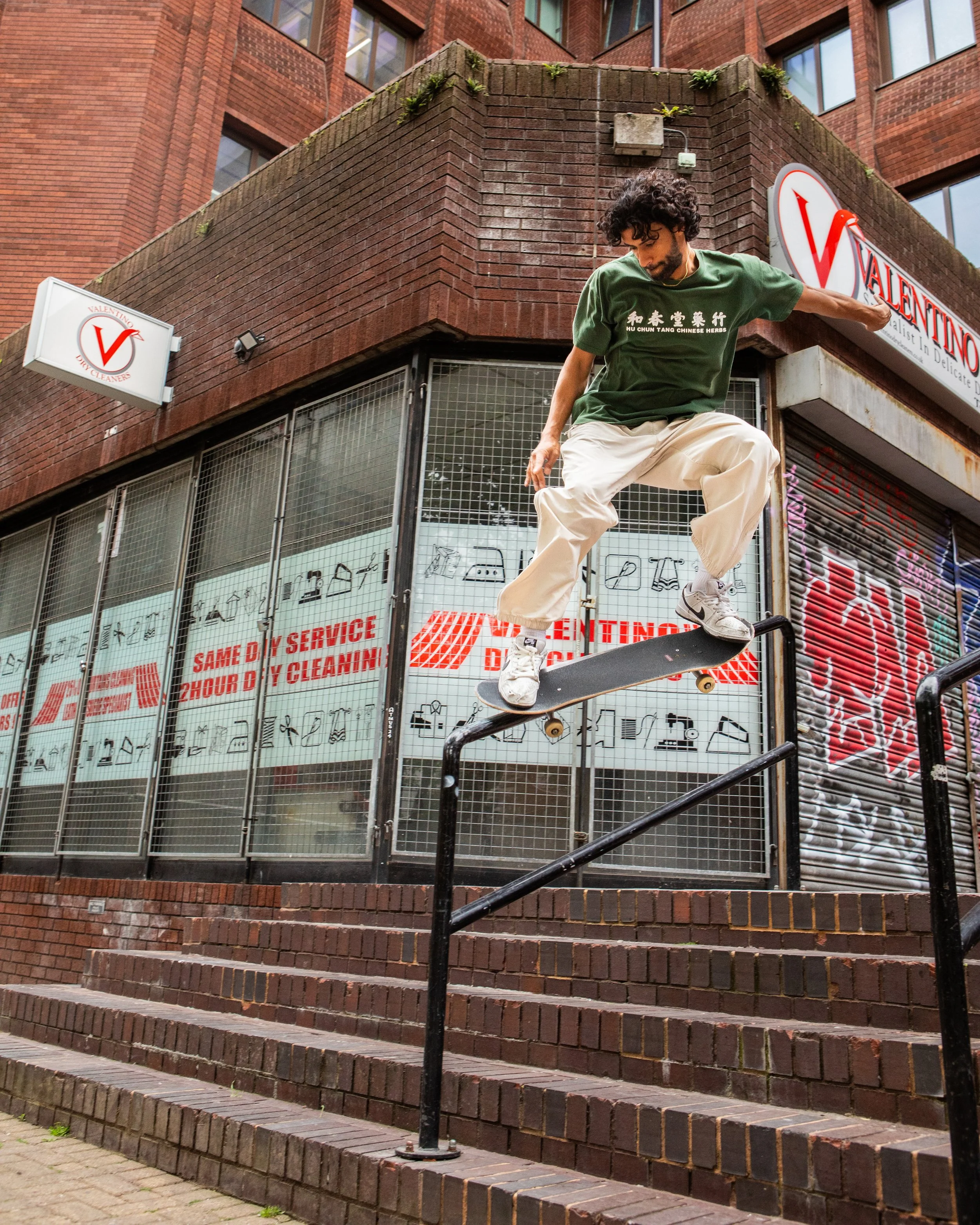 A man skateboarding on a rail outside a building with a sign that says Valentino Dry Cleaners.
