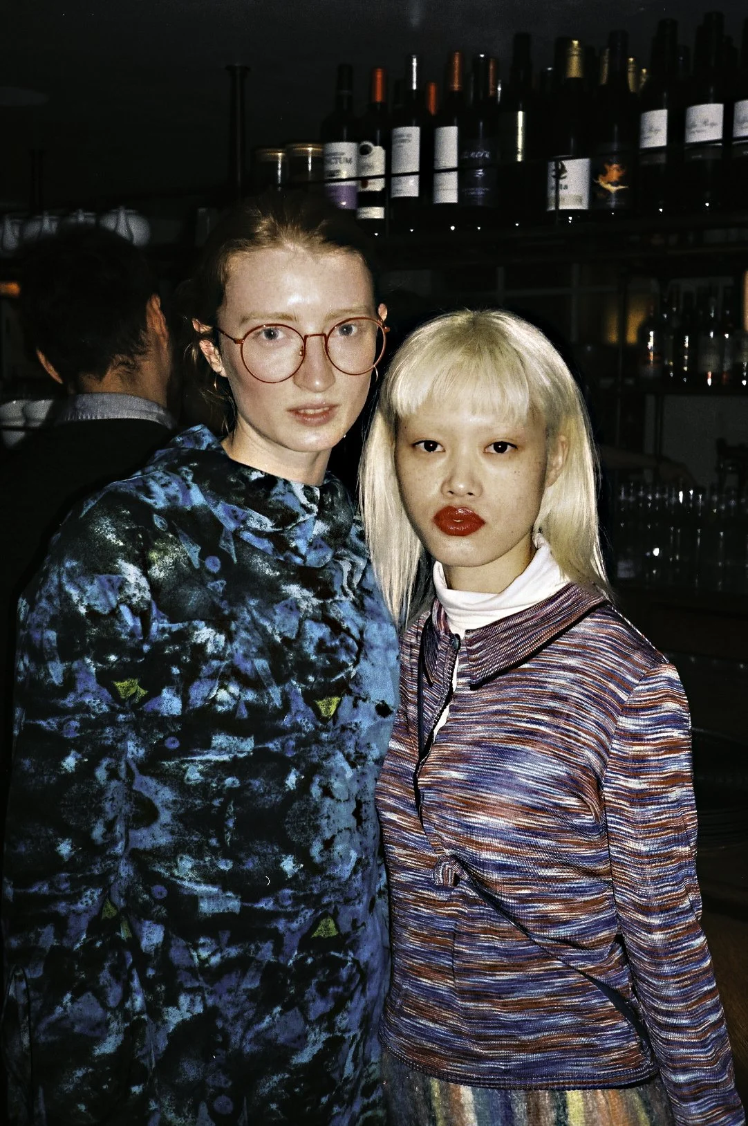 Two women standing close together in a bar or restaurant, with shelves of wine bottles in the background. The woman on the left has light skin, red hair, glasses, and is wearing a blue and black patterned top. The woman on the right has light skin, platinum blonde hair, red lipstick, and is wearing a multicolored striped jacket over a white turtleneck.
