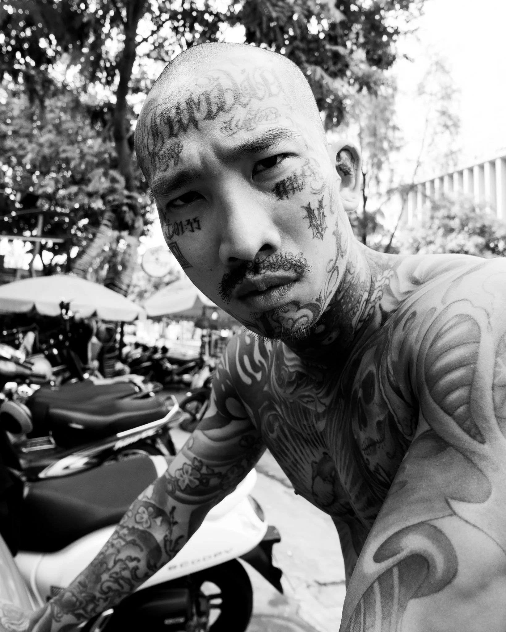 Black and white close-up photo of a tattooed man with a shaved head, narrow eyes, and a mustache, outdoors with trees, buildings, motorcycles, and umbrellas in the background.