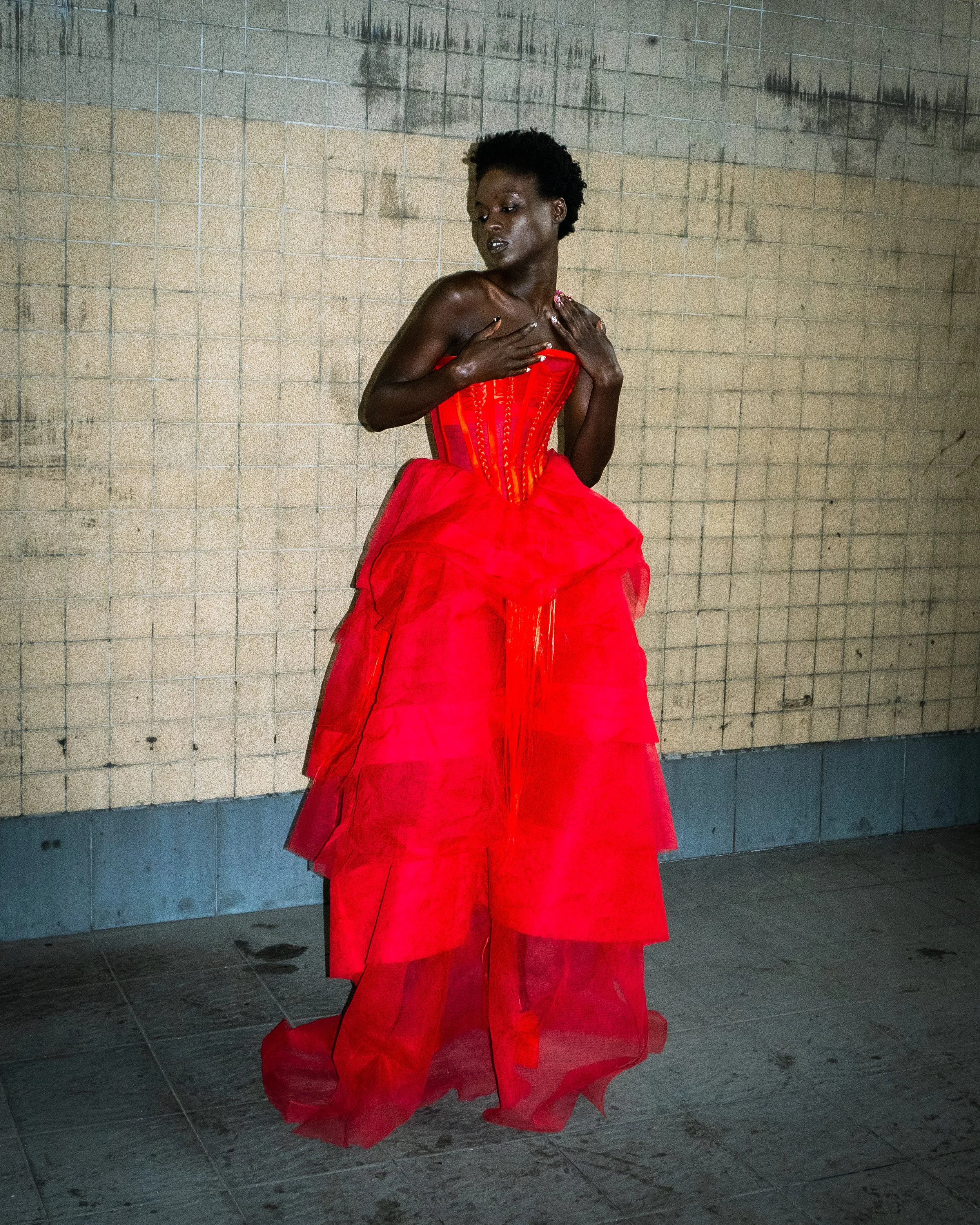 A woman wearing a bright red gown with a corset top and layered tulle skirt, standing against a beige tiled wall, with her hands on her chest and her eyes closed.