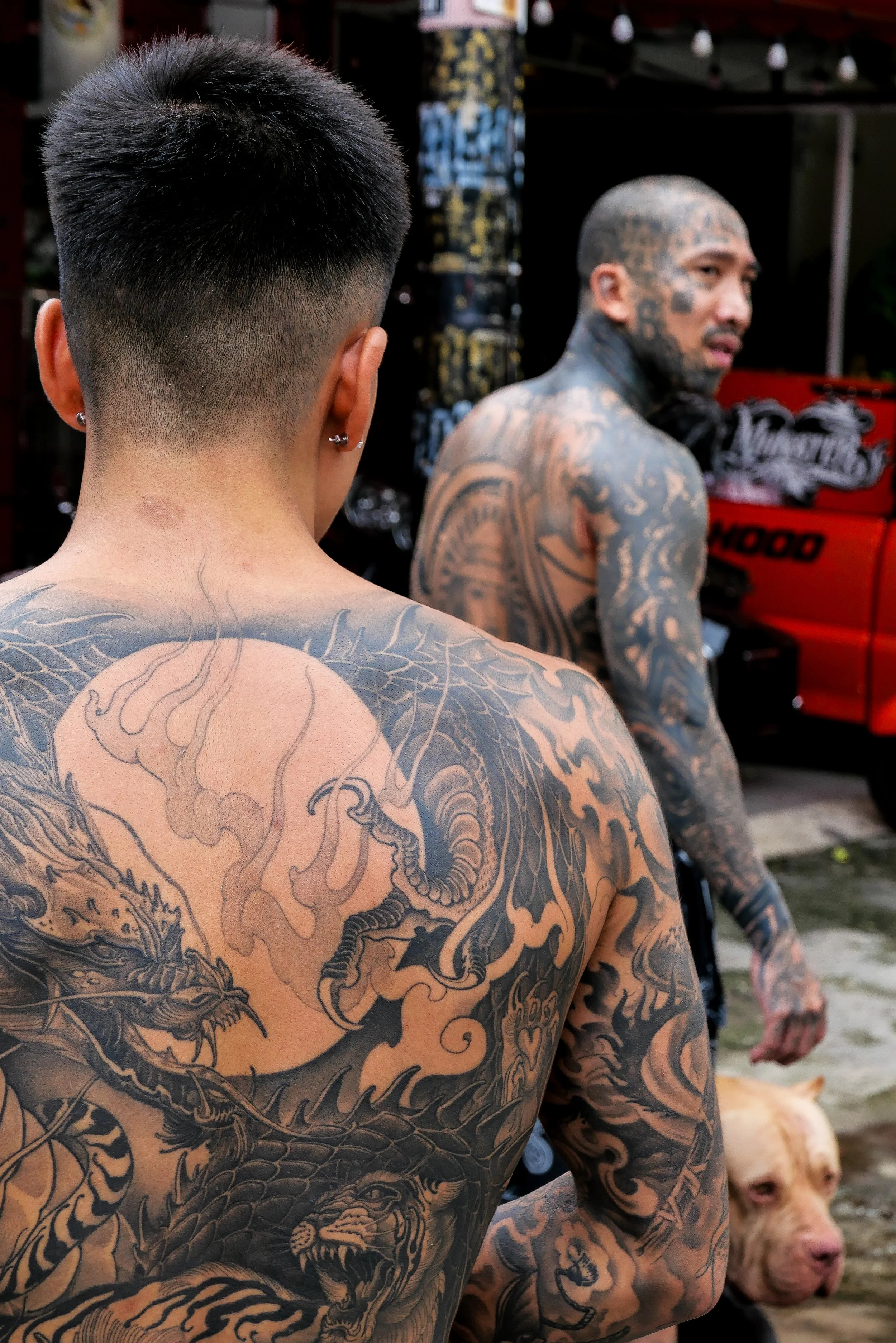 Two tattooed men, one with a fade haircut and earrings facing away, and the other with a full head of tattoos turned to the side, standing outdoors near a red cart and a dog.