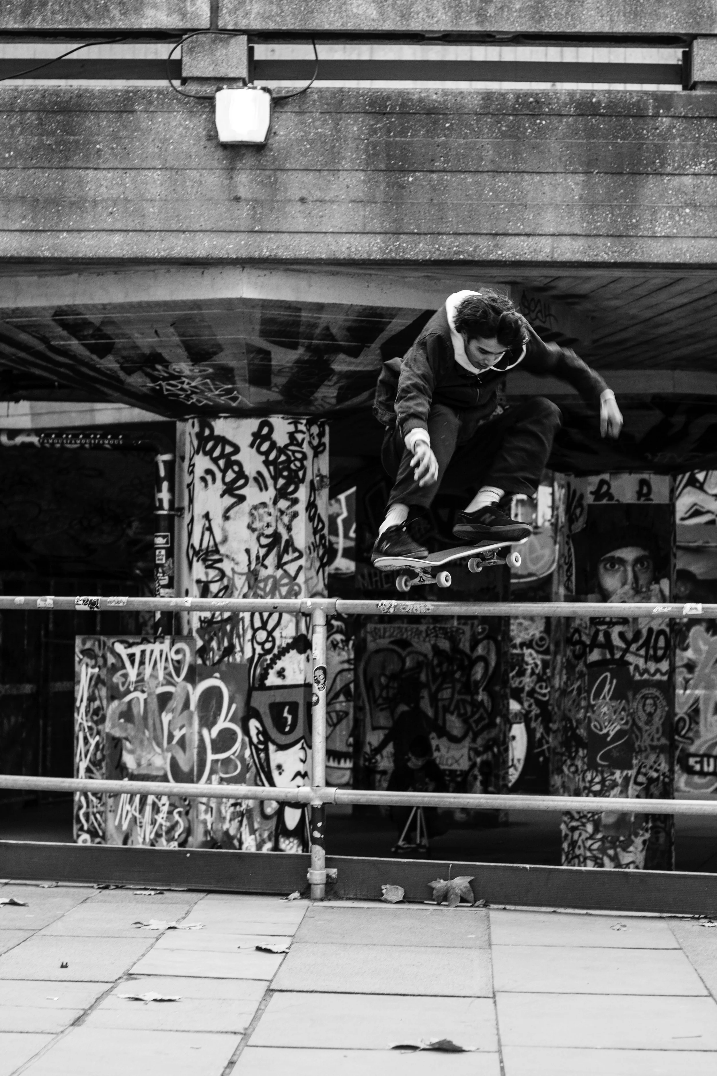 A person skateboarding on a rail under a graffiti-covered bridge.