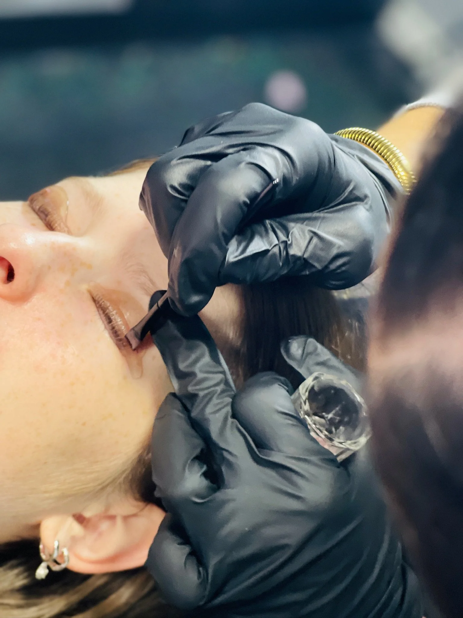 A person lying down with their eyes closed, receiving a facial treatment or medical procedure, wearing black gloves and a black jacket, with a glass of water nearby.