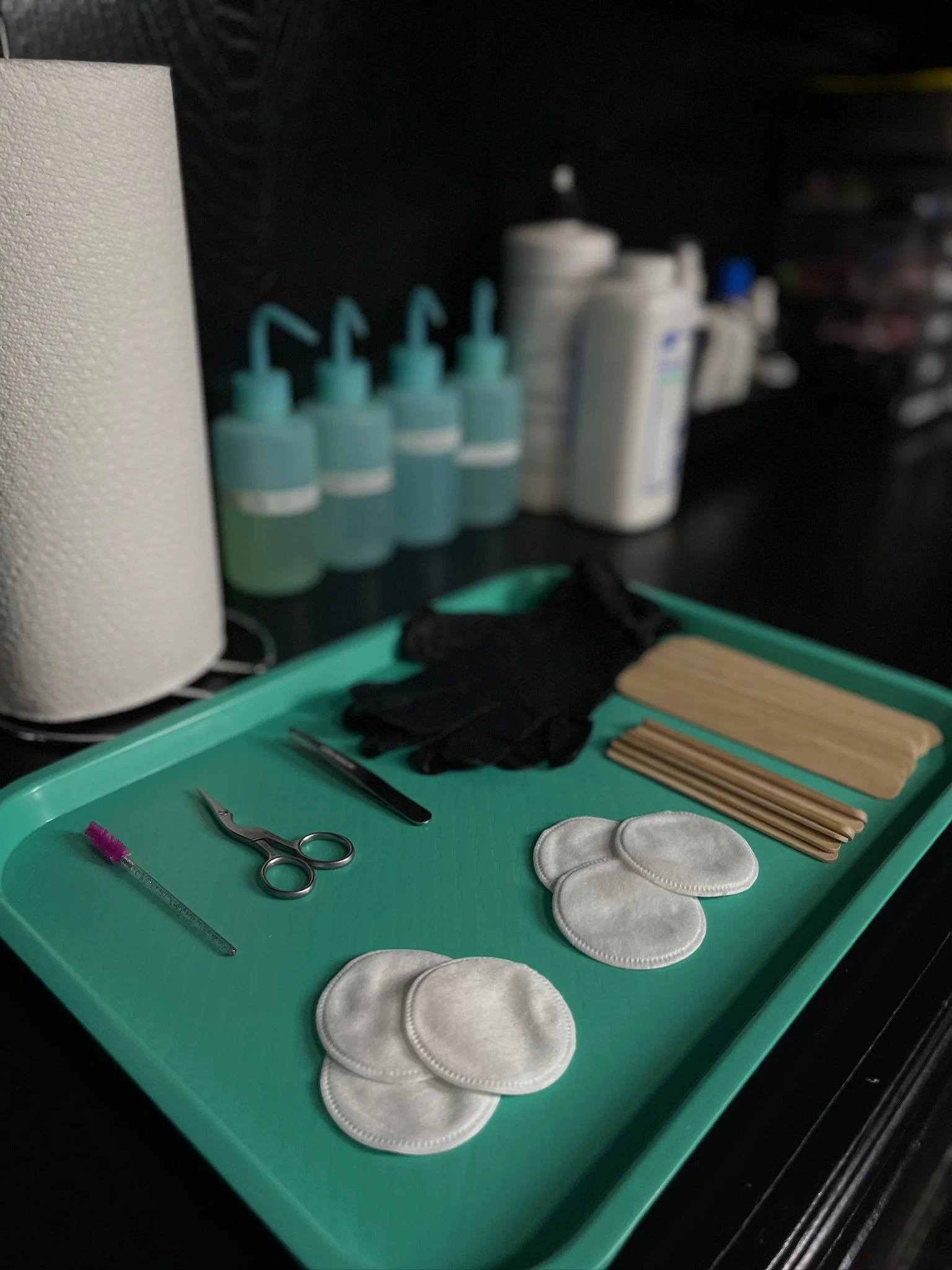 A teal-colored tray holds various items including white cotton pads, black gloves, wooden tongue depressors, metal scissors, a small pink applicator, and a pair of tweezers, with a workspace visible in the background.