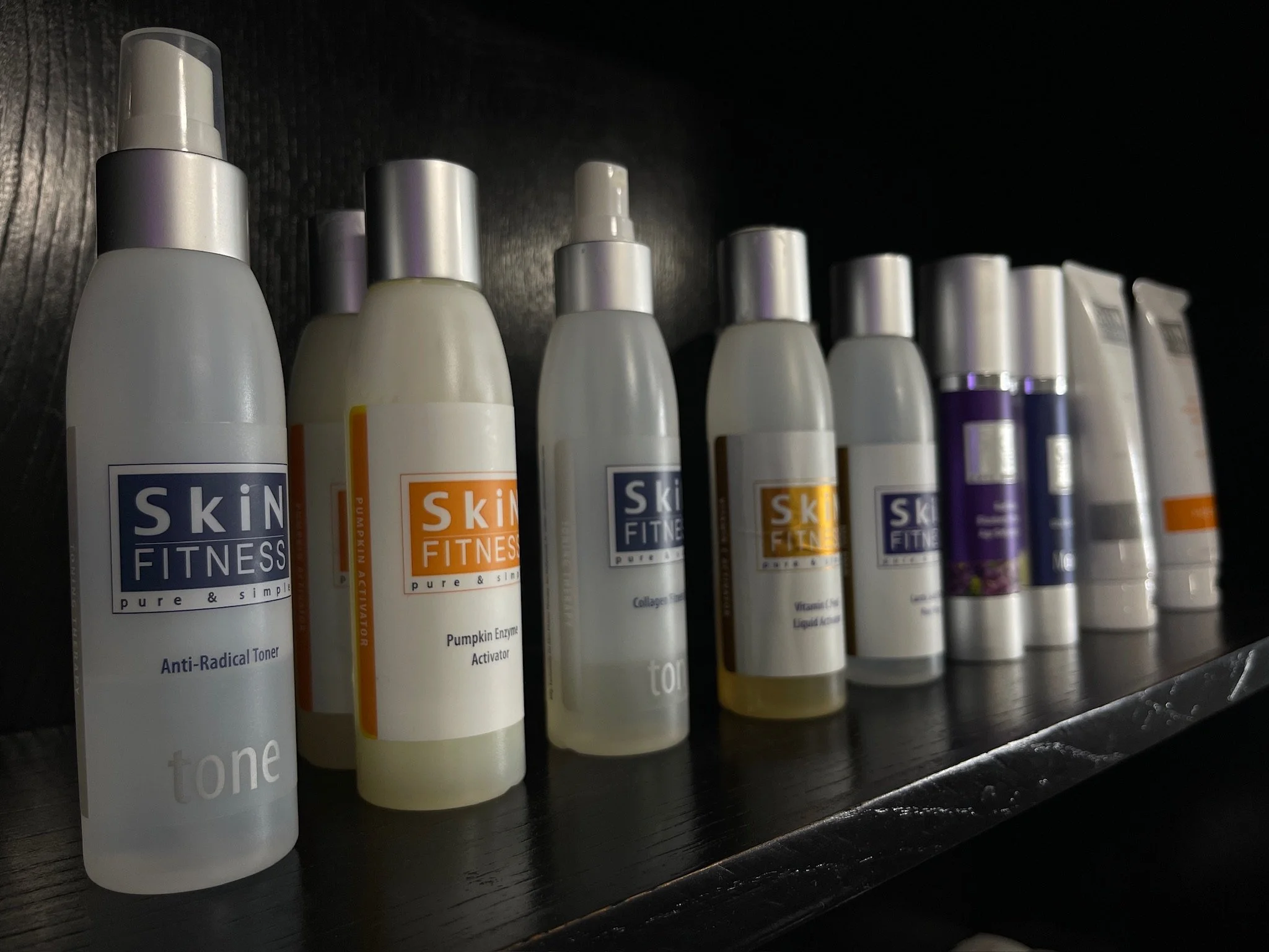 A row of skincare bottles on a black shelf, including toner, pumpkin enzyme activator, collagen, vitamin C, and other skincare products, some with purple accents.