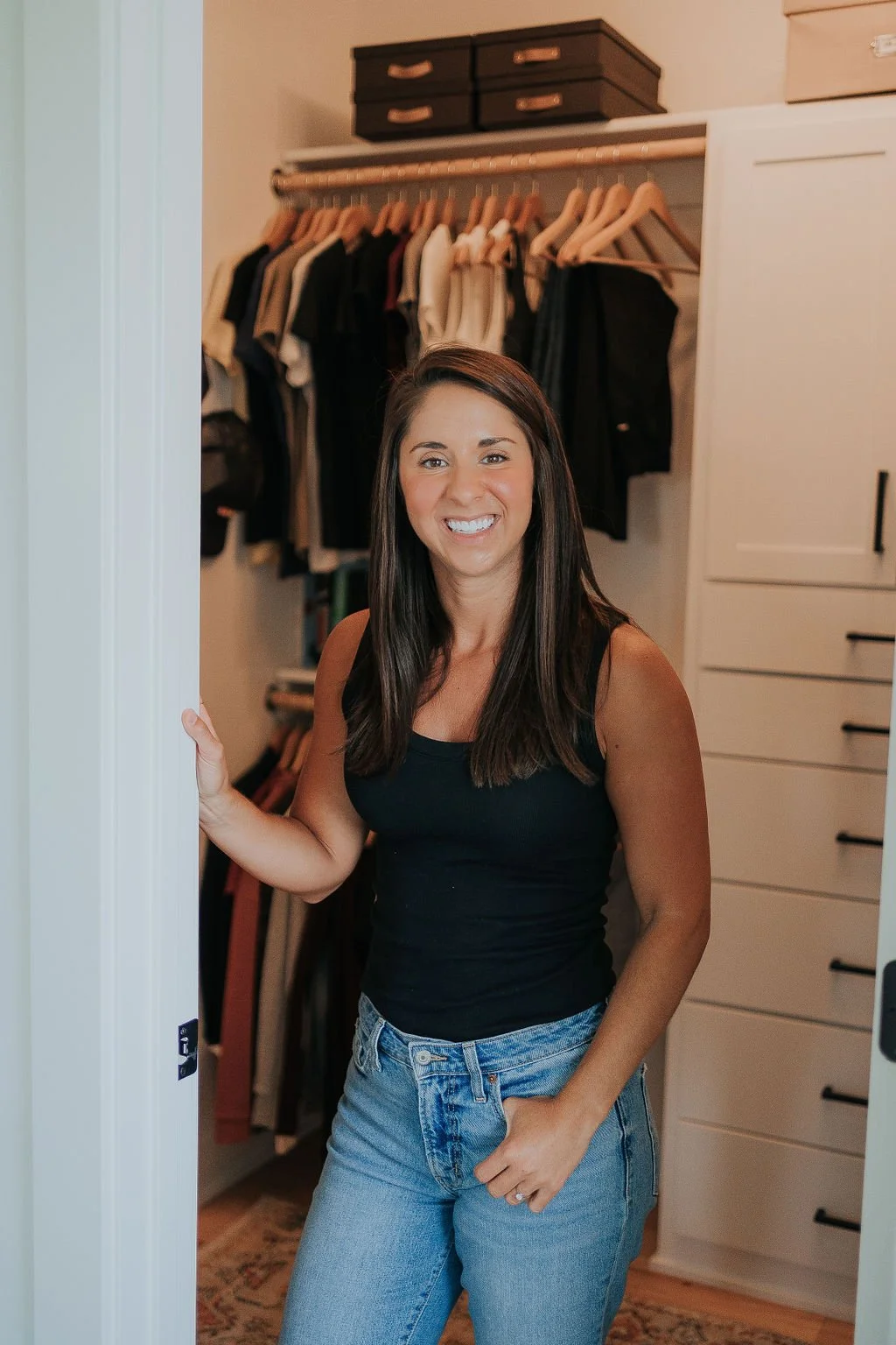 Professional organizer for ADHD, Melissa Welch, smiles and leans against the wall of her organized, clutter-free closet that includes easy visibility for neurodivergence, hooks, and easily grouped items.