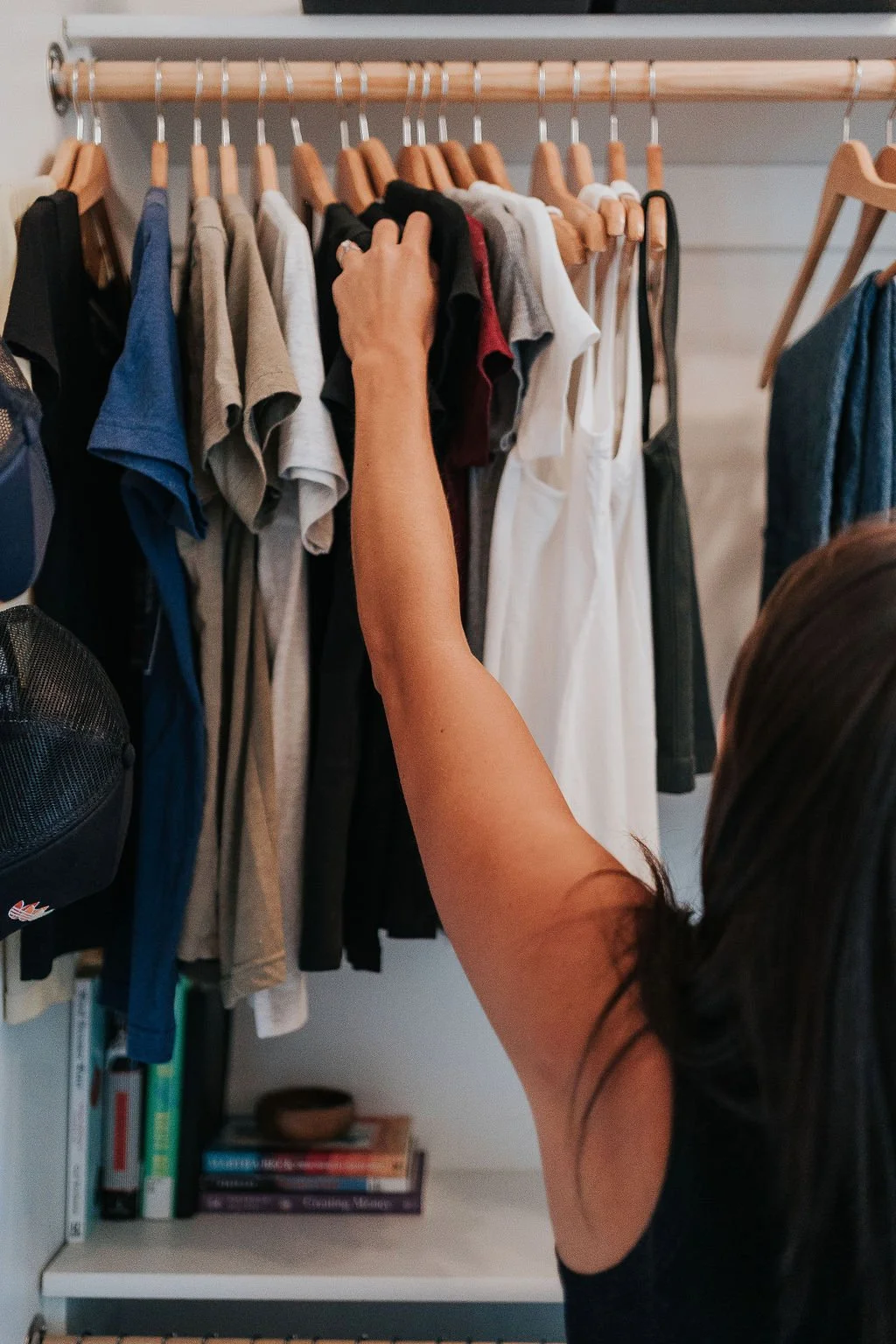 Melissa Welch, an organizer for ADHD, hangs a black tee shirt in an organized closet. Hats hang on hooks beside her and library books are lined on the shelf.