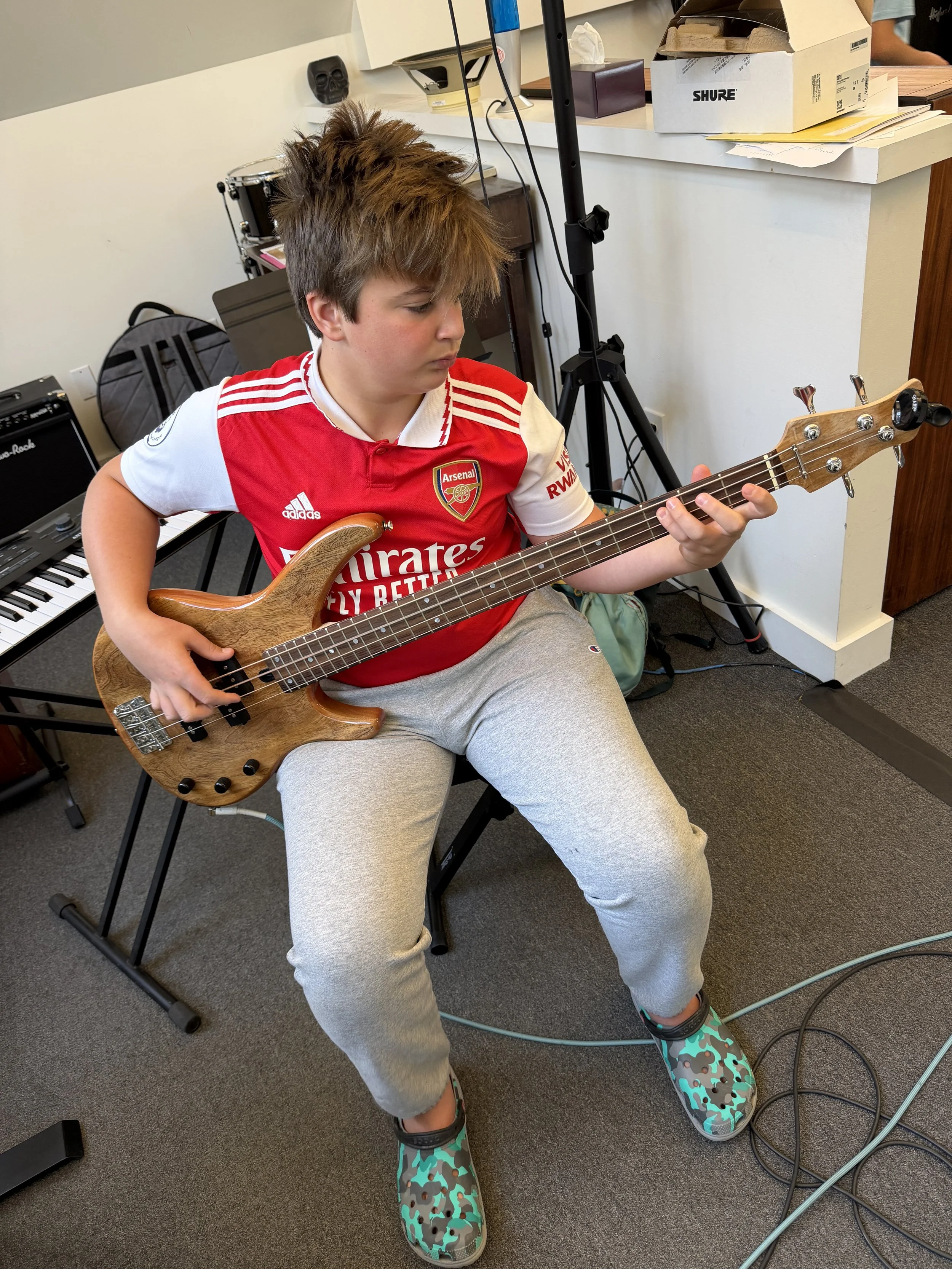 A young boy sitting on a chair, playing a wooden electric bass guitar. He is wearing a red Arsenal football jersey, gray sweatpants, and colorful slip-on shoes. The background shows musical equipment and various boxes on a counter.