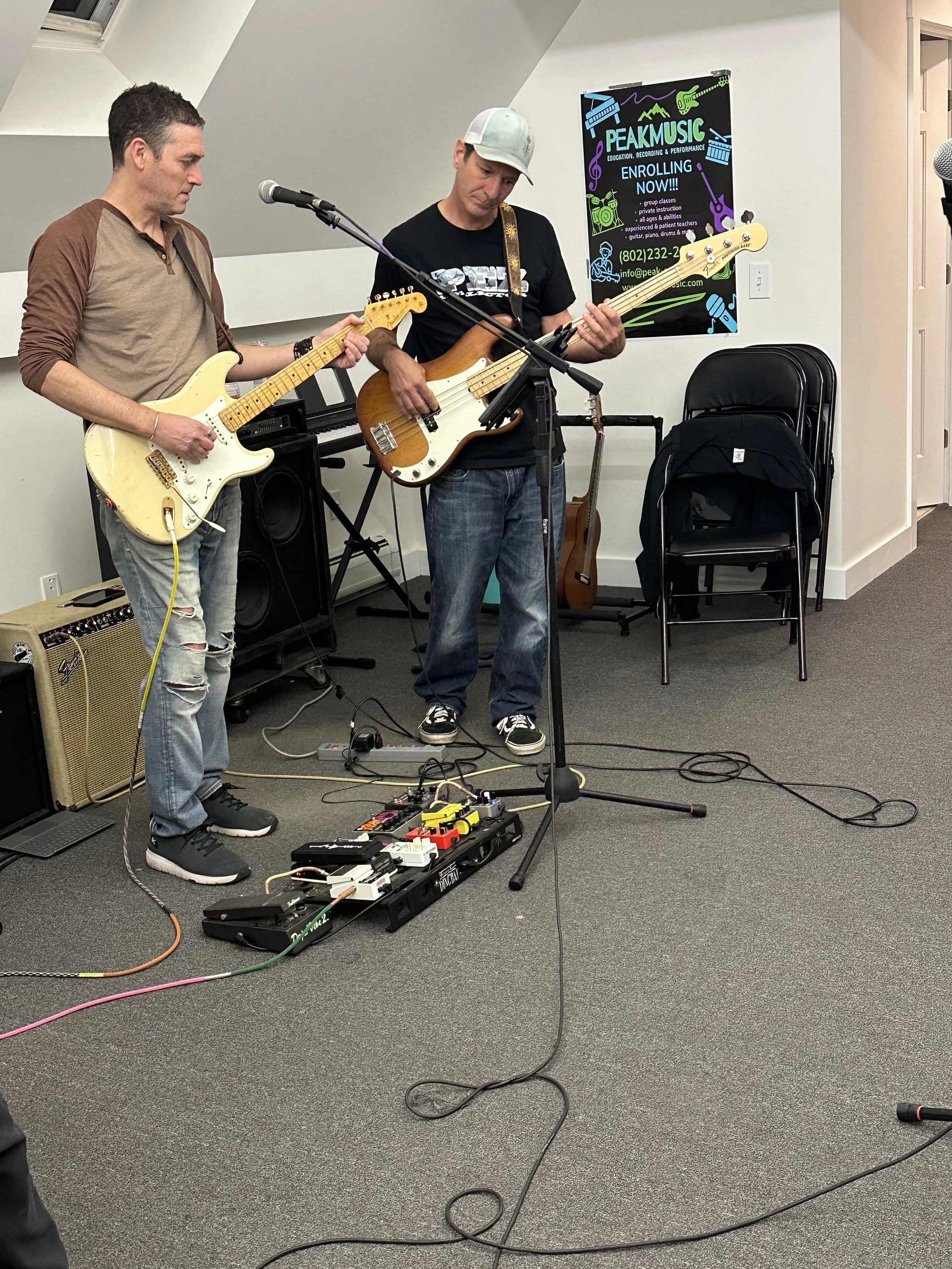 Two men playing electric guitars in a room, with musical equipment on the floor and a banner in the background advertising Peak Music classes.